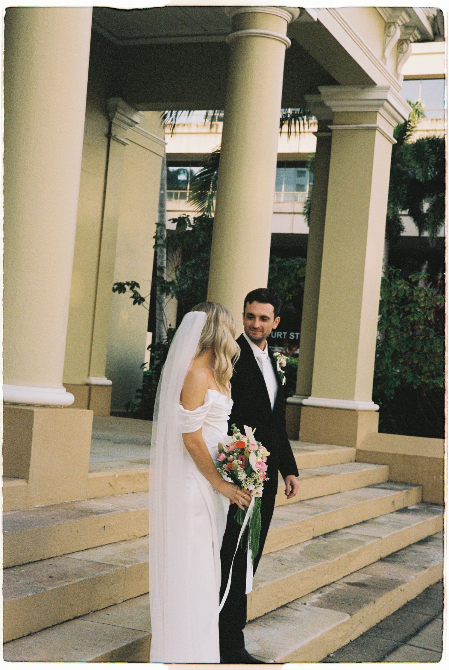 Bride in white off-shoulder gown with veil and bouquet stands with groom in black suit on steps of elegant venue.