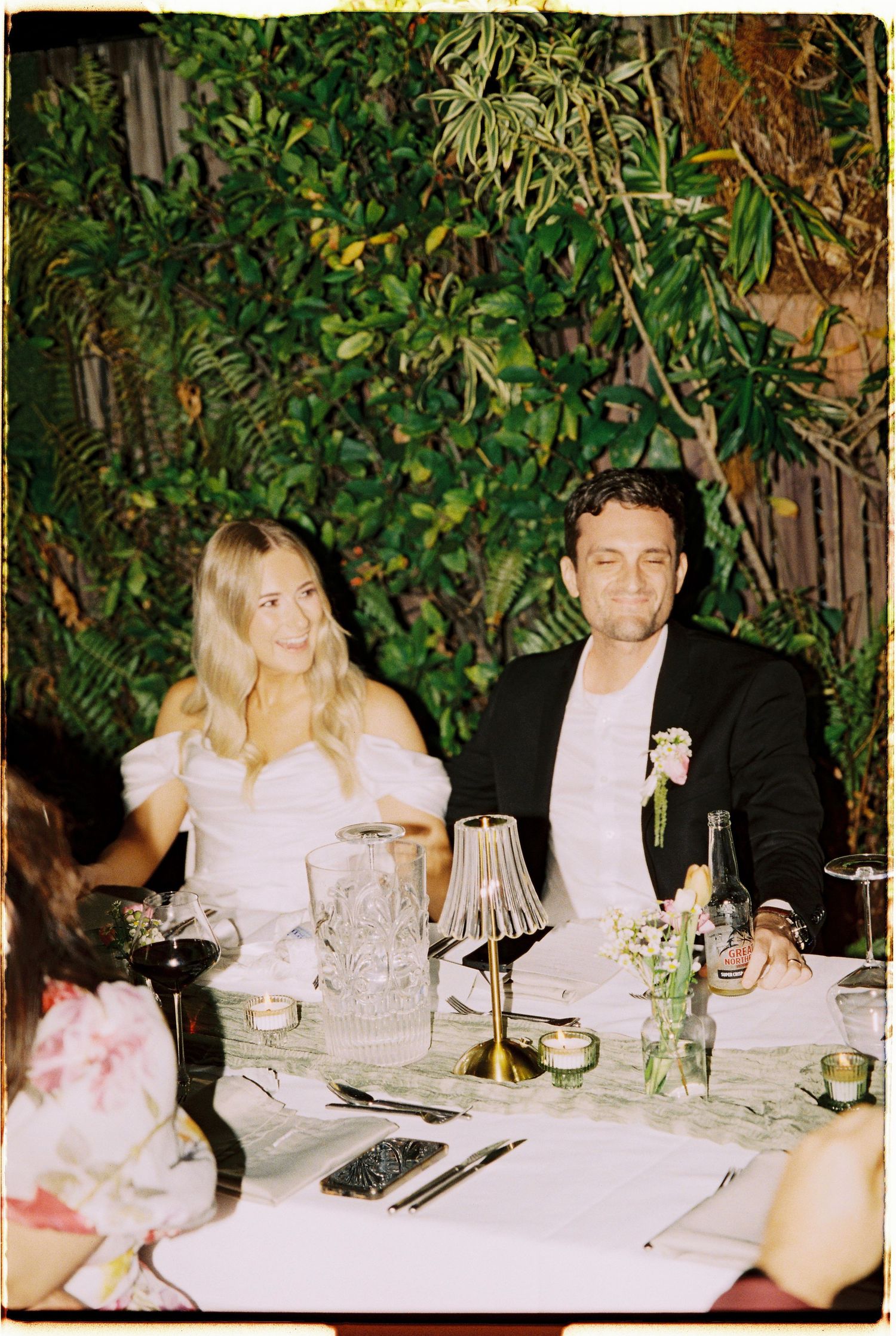 A blonde bride in a white off-shoulder dress sits beside her groom in a black suit at their wedding reception table.