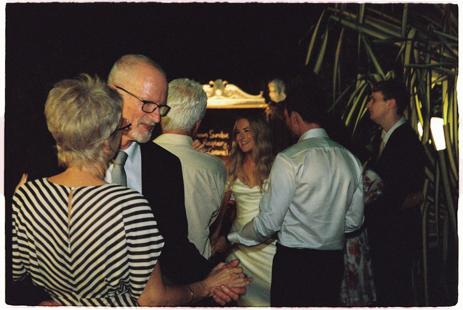 Guests mingling at an elegant outdoor wedding reception at night, with bride in white dress greeting attendees.