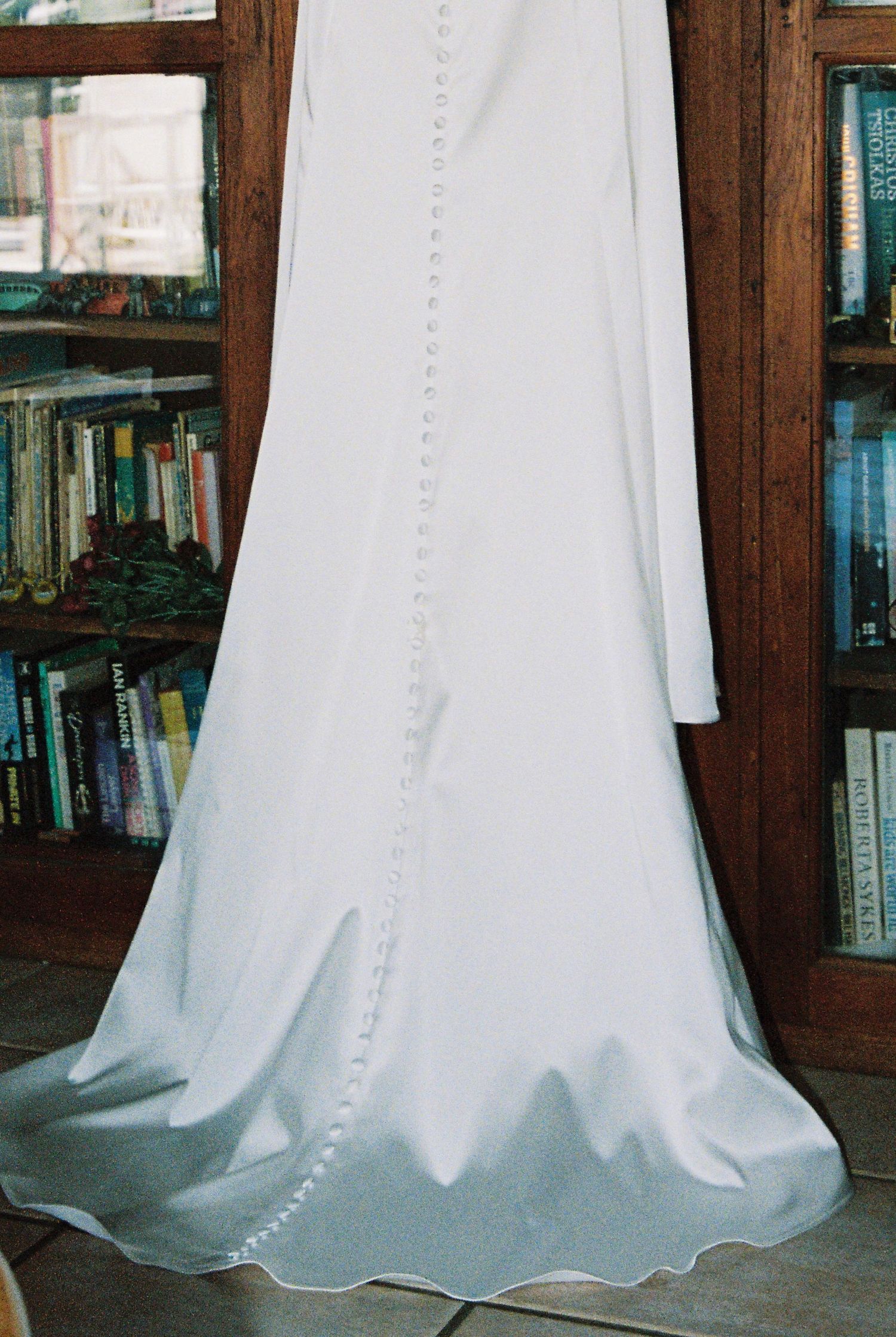 White wedding gown with button back detail hanging near wooden bookcase.