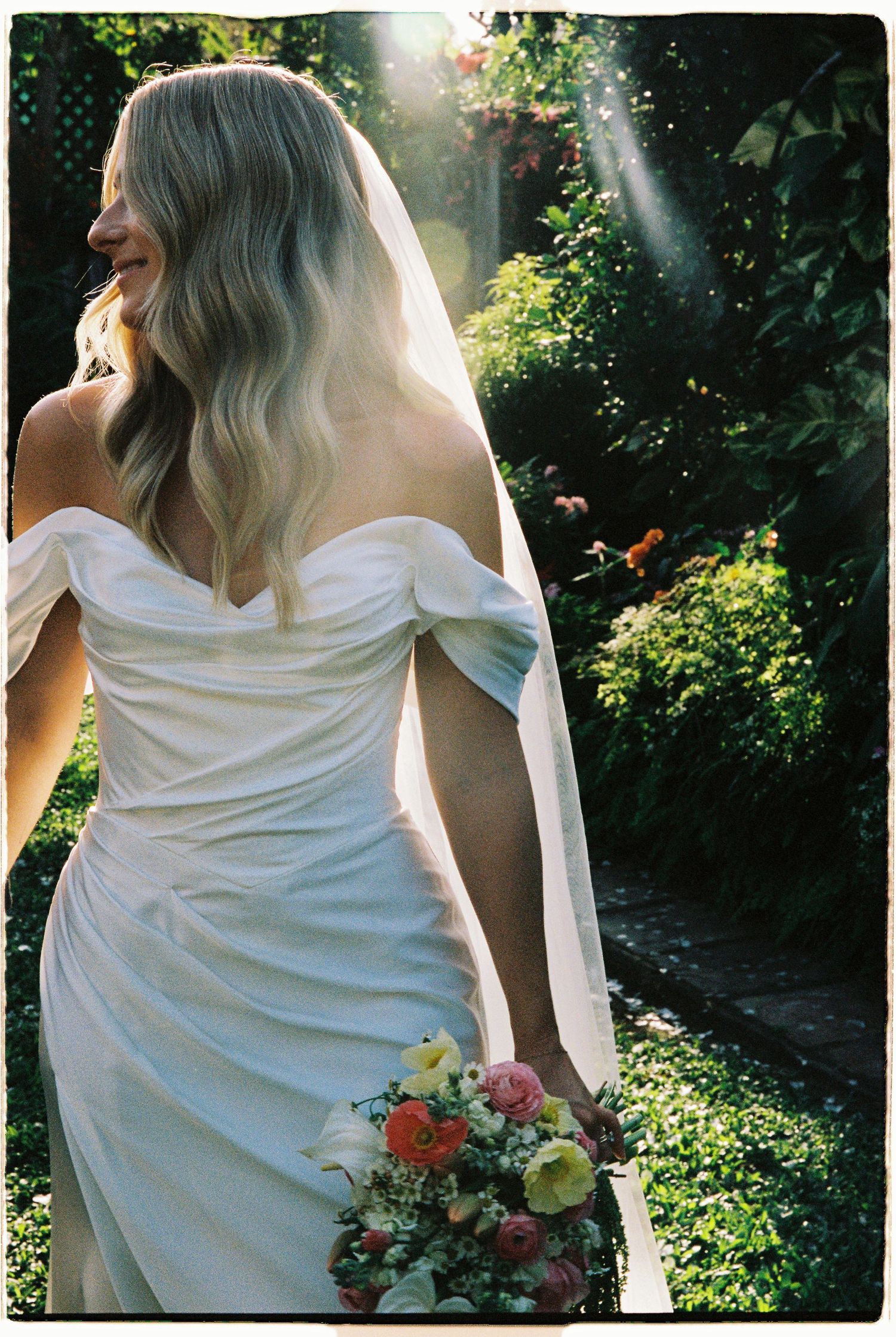 Bride in off-shoulder white gown holds colorful bouquet in sunlit garden.