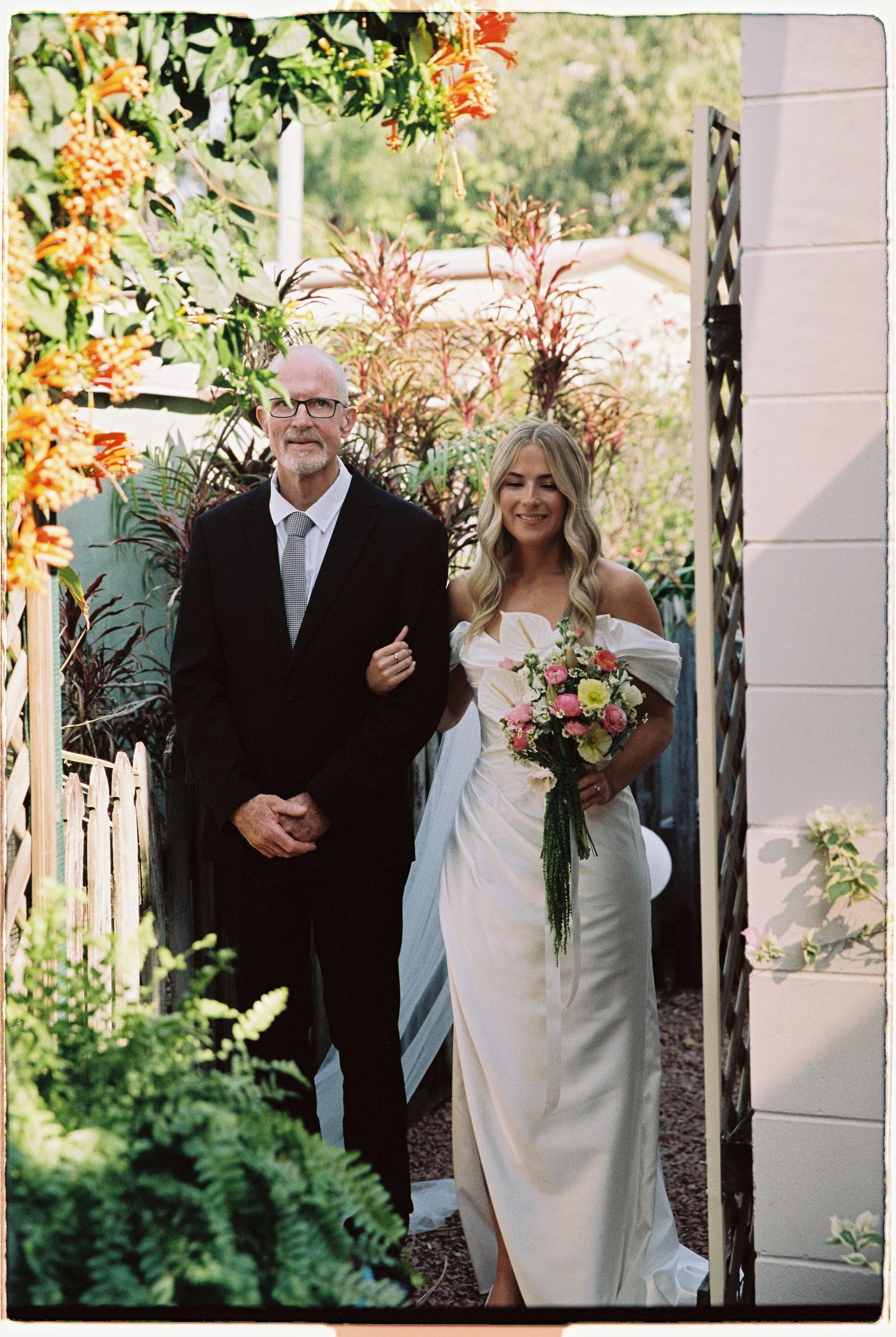 Bride in elegant white dress holds colorful bouquet while walking with older gentleman through garden gate.