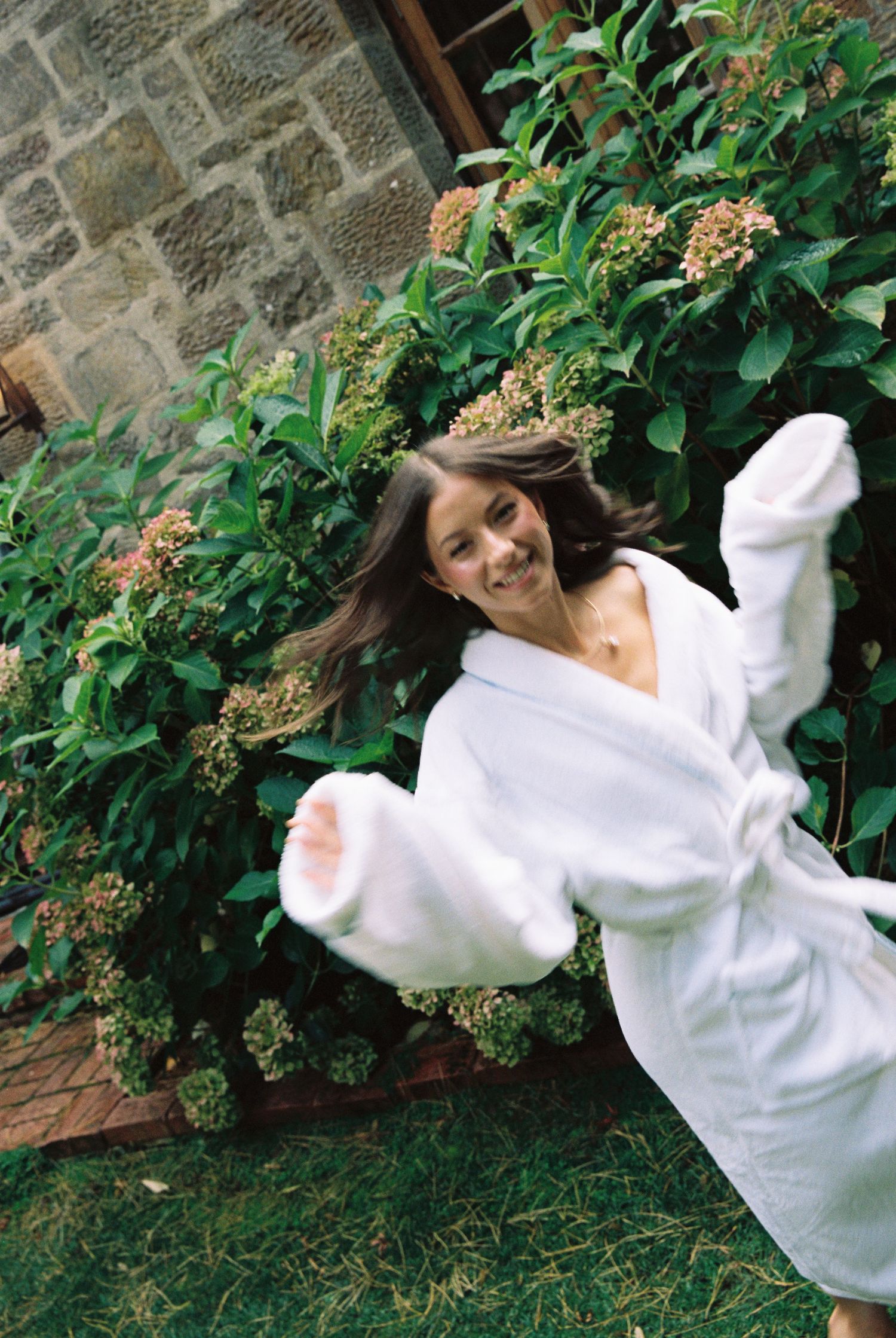 Joyful bride in a white robe twirling in a garden surrounded by blooming hydrangea bushes.