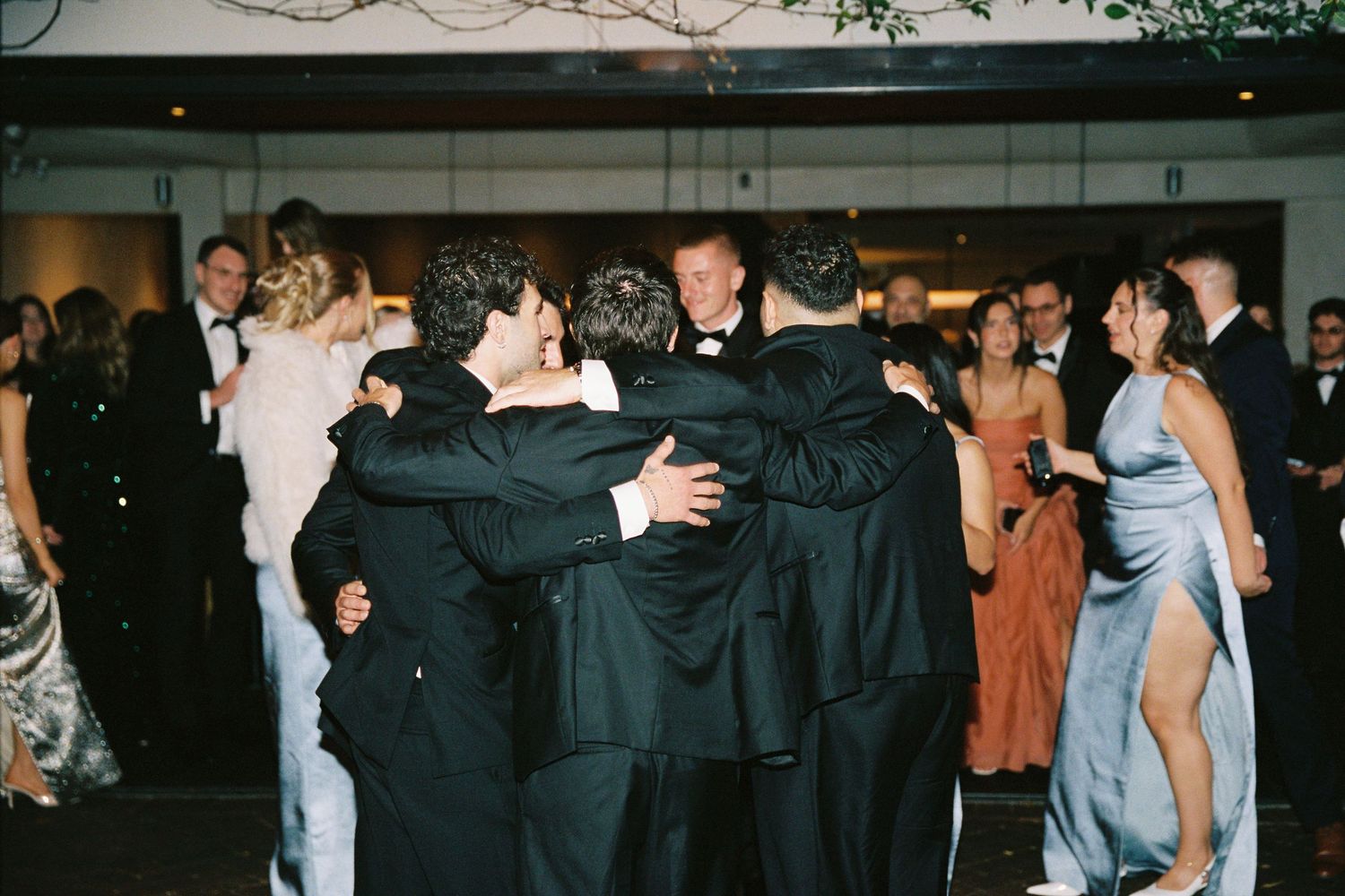 Wedding guests in formal wear share a group hug on the dance floor.