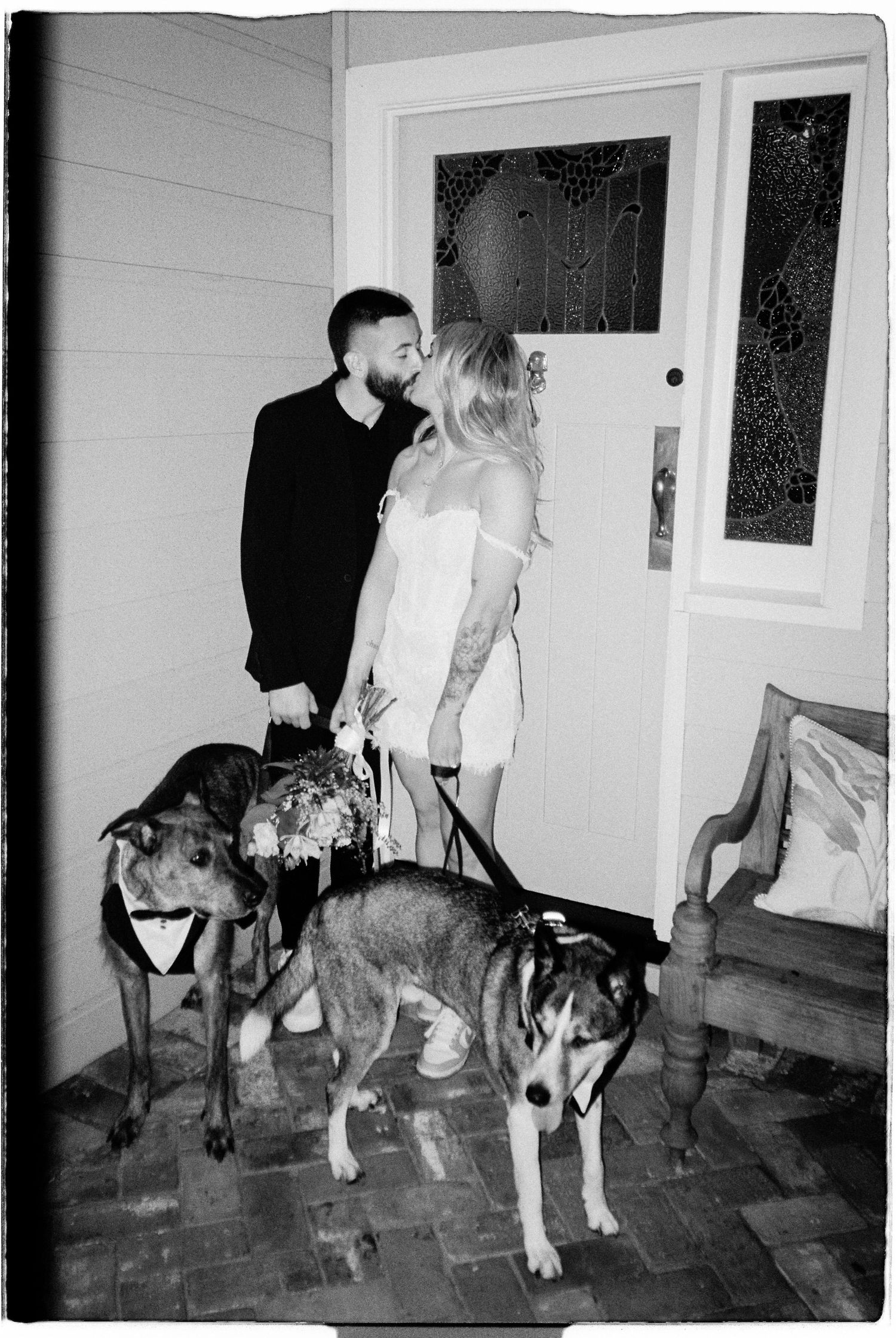 Black and white photo of a couple kissing in wedding attire with two dogs wearing bow ties at their front door.