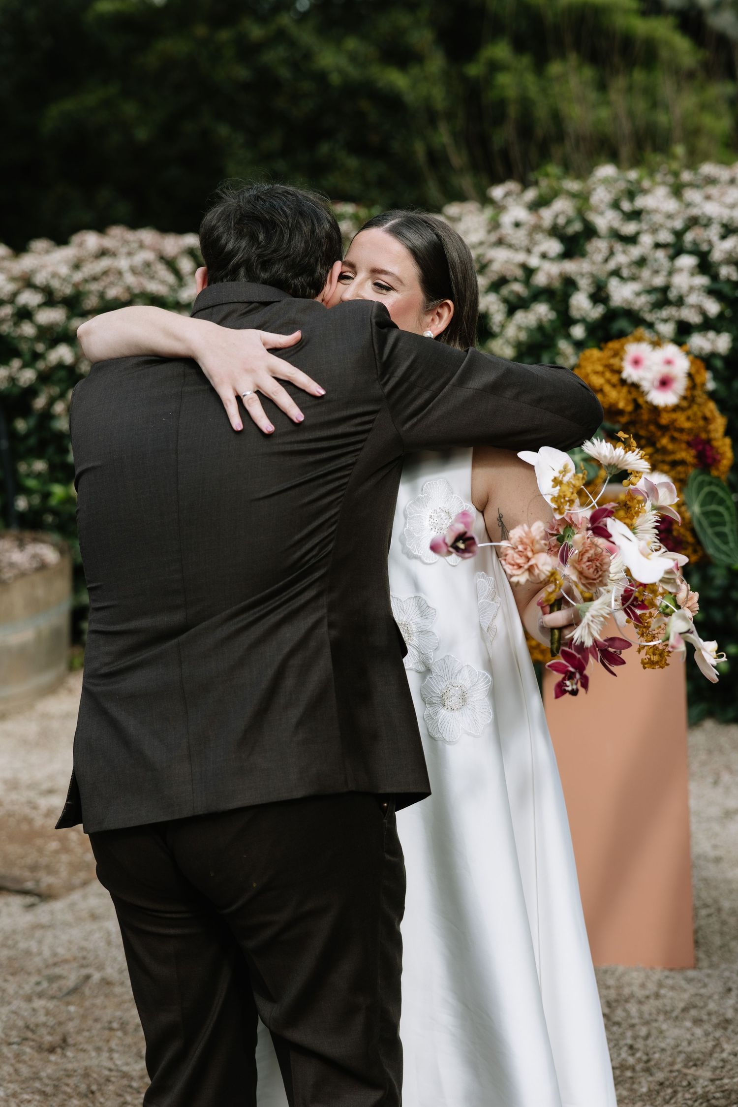 Bride in white gown hugging a man in black suit, holding colorful bouquet, with flower garden backdrop.