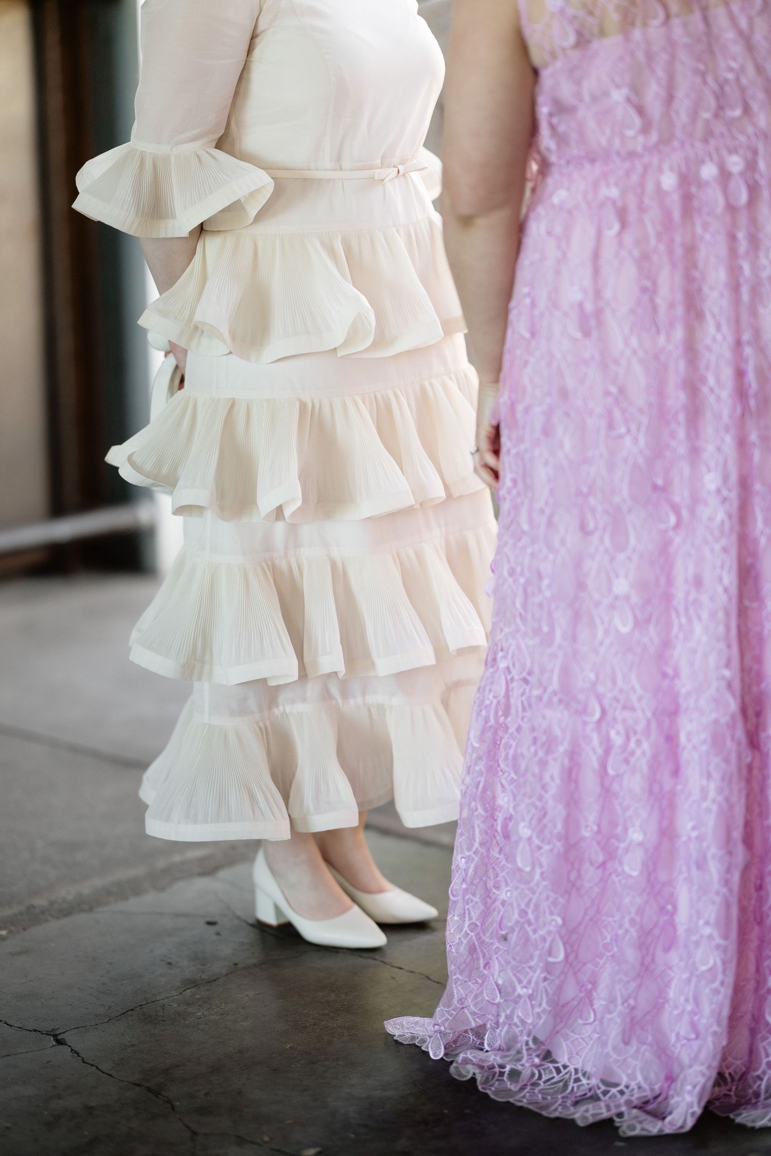 Detail shot of bride in ivory ruffled tiered dress standing beside guest in pink lace gown.