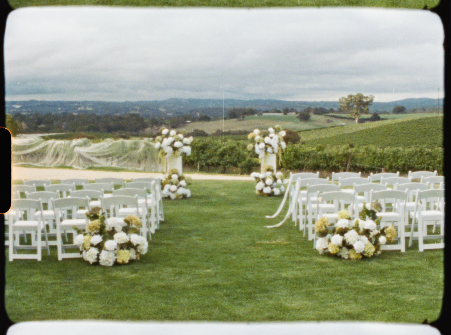 Elegant outdoor vineyard wedding ceremony setup with white chairs and white floral arrangements.