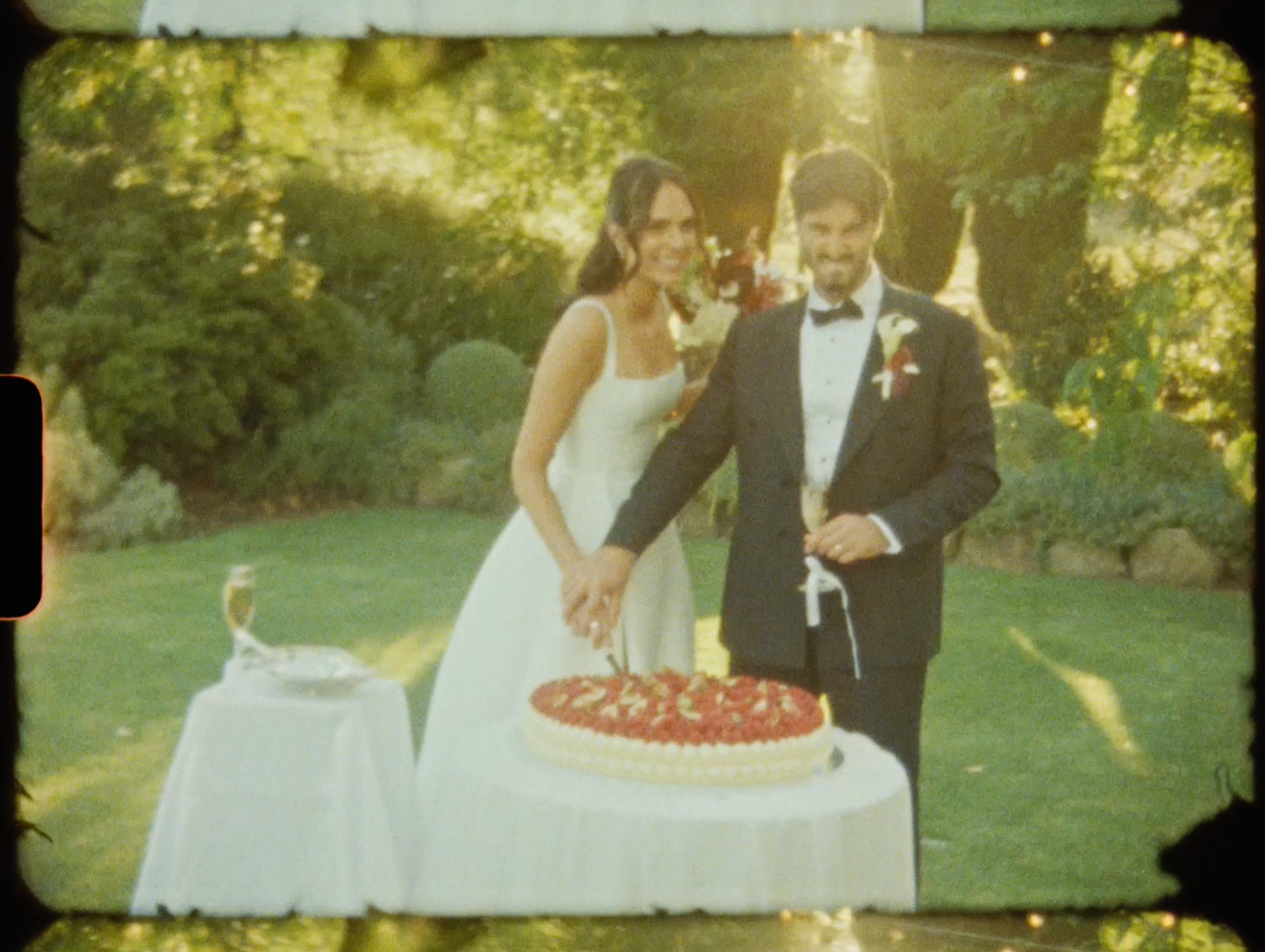 Bride and groom cutting their strawberry wedding cake together outdoors in a garden setting.