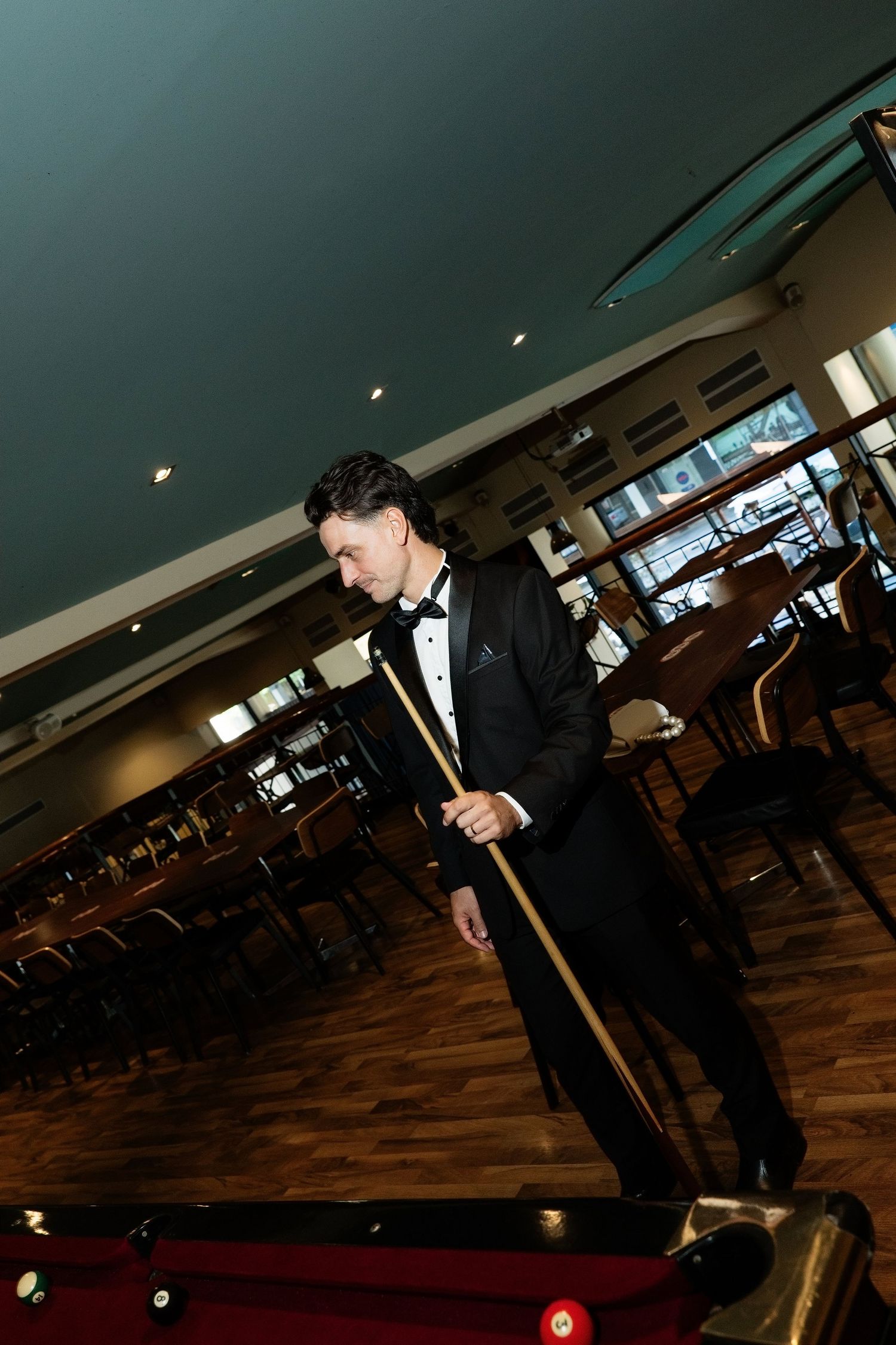 Groom in black tuxedo holding a pool cue while standing beside a red billiard table in a venue.