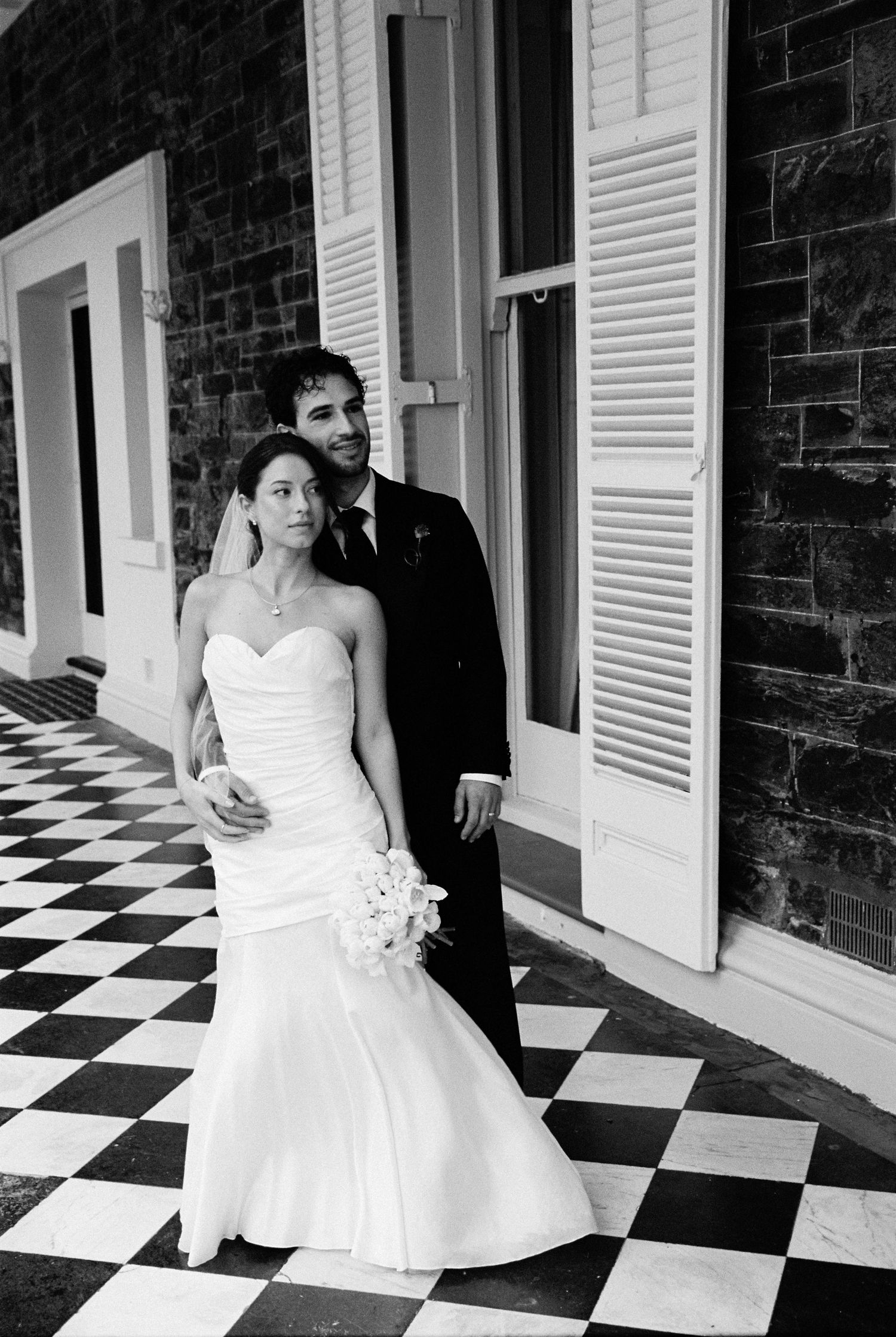 Bride and groom posing on checkered floor beside brick wall with louvered shutters.