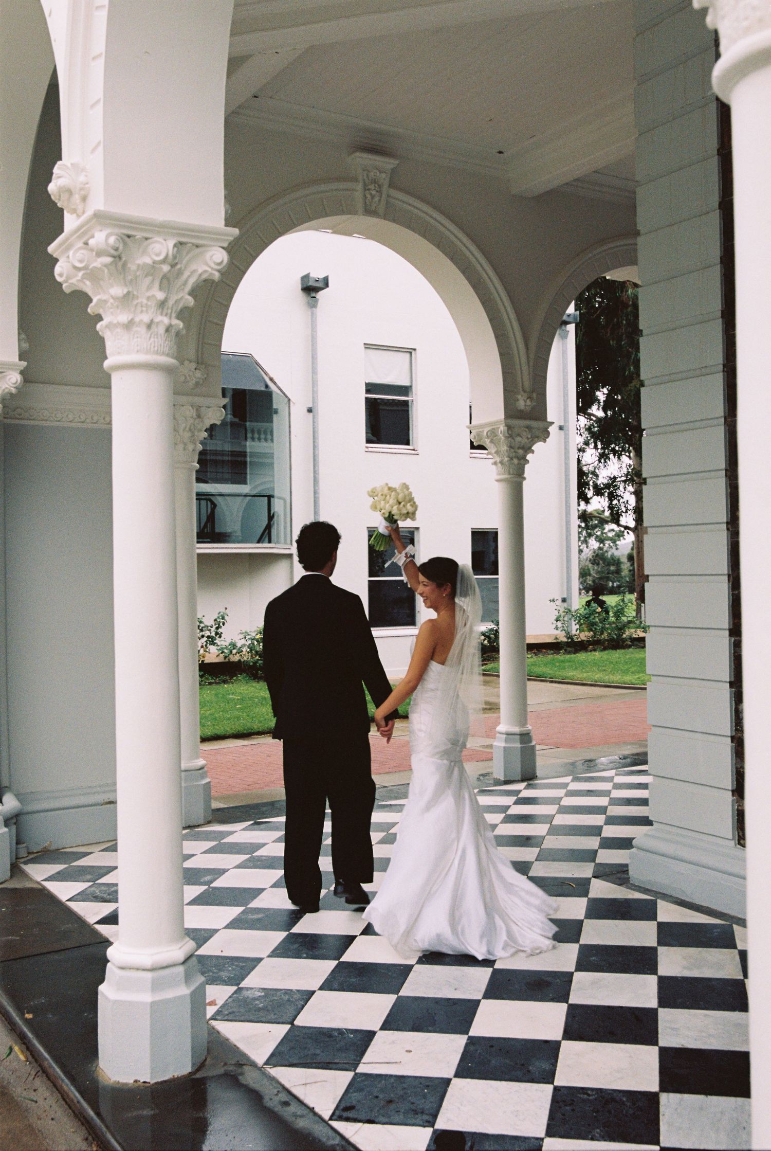 Bride in white gown and groom in black suit hold hands under elegant white arches on checkered floor.