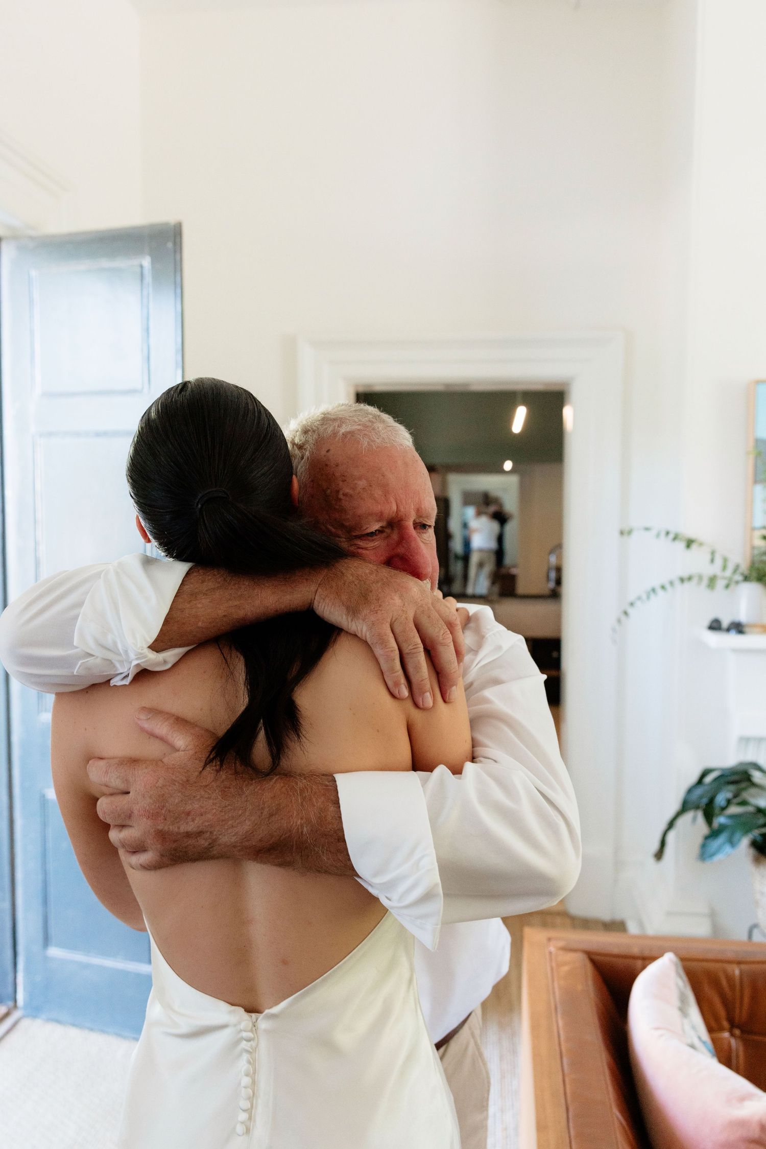 Emotional father hugging bride in backless white wedding dress during heartfelt first look moment.
