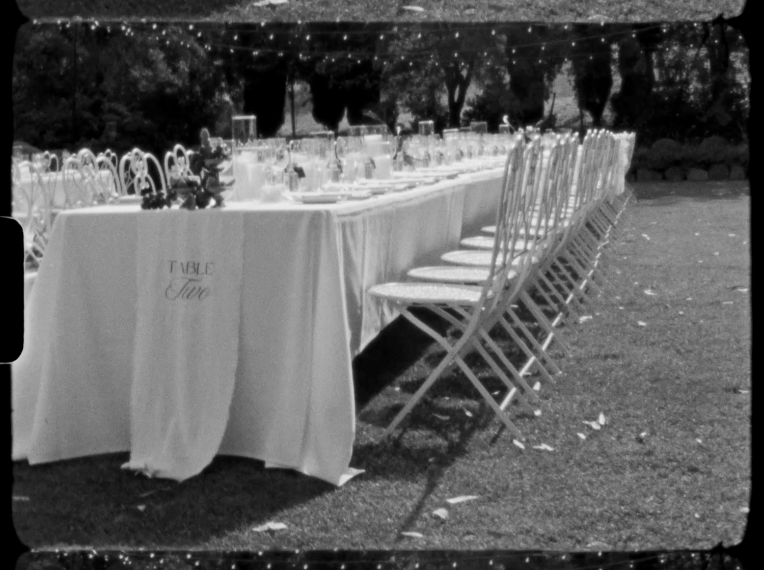 Black and white photo of a long elegantly set outdoor banquet table with white linens and folding chairs on a lawn.