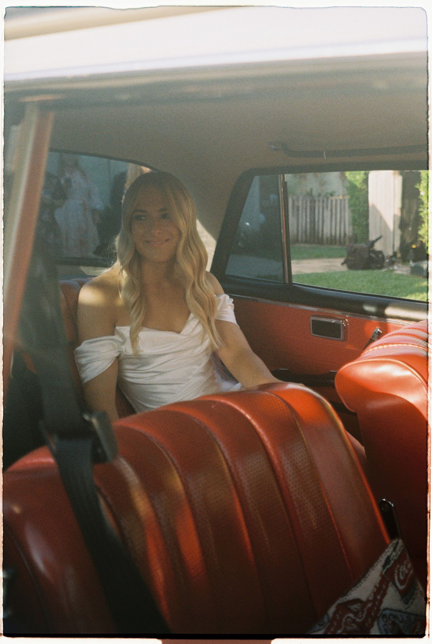 Blonde bride in white off-shoulder dress sitting in vintage car with red leather interior.