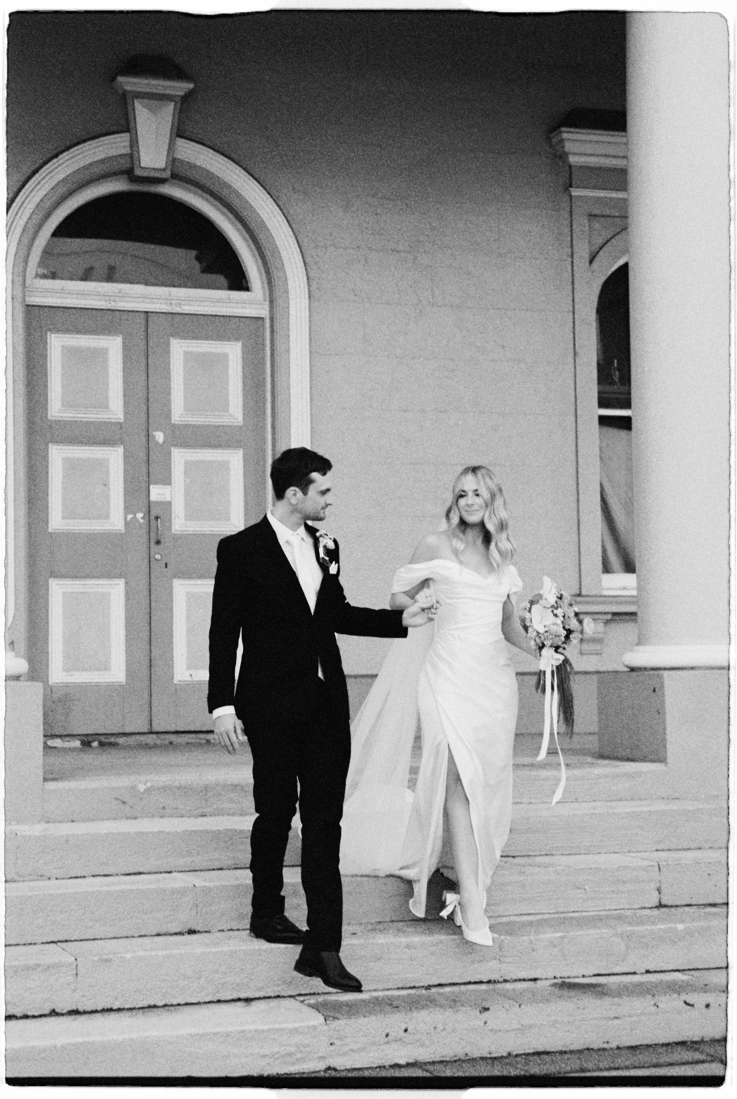 Newlywed couple walks down church steps, bride in off-shoulder gown holding bouquet, groom in black tuxedo.