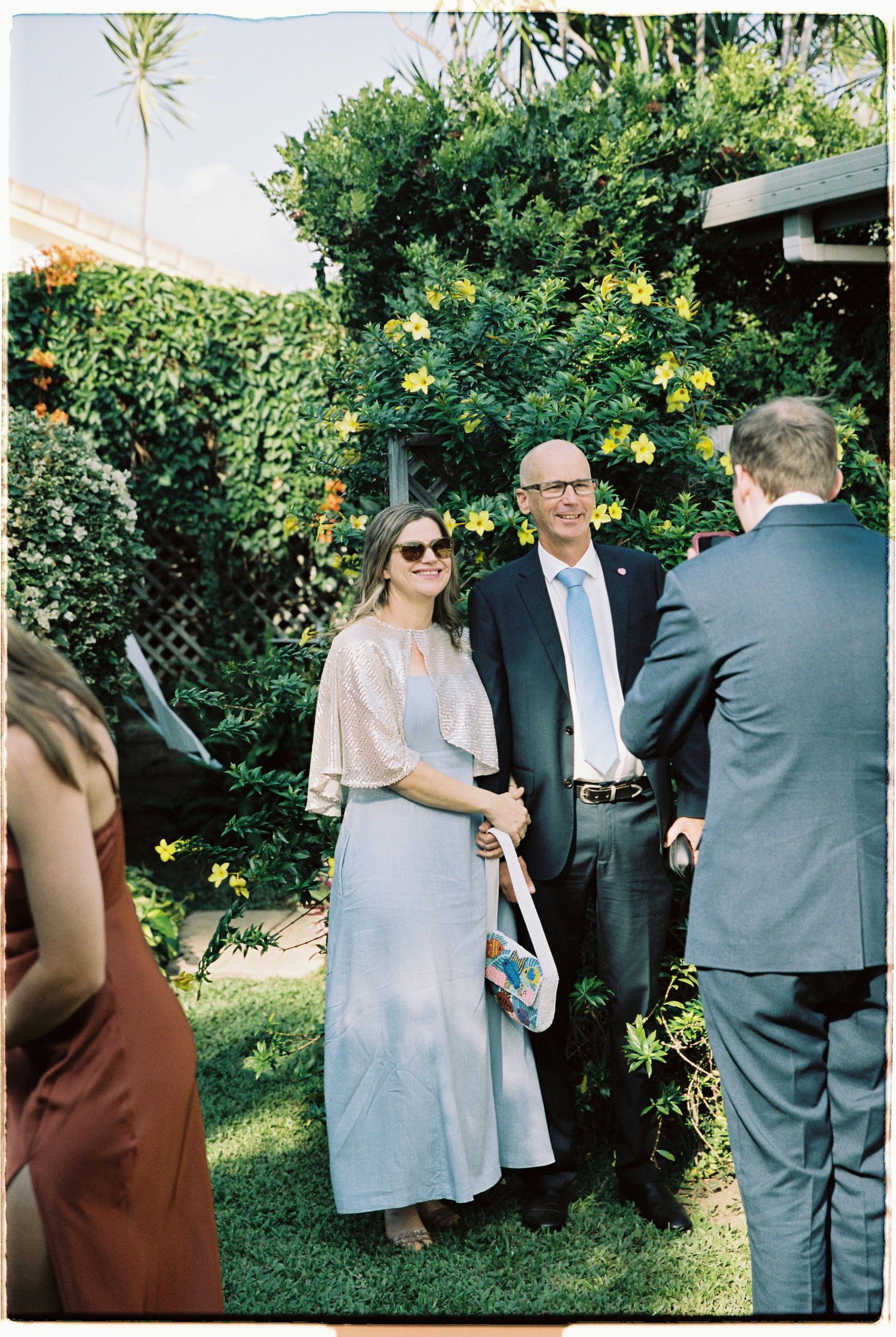Wedding guests in garden venue, woman in silver dress with sunglasses greeting suited man.