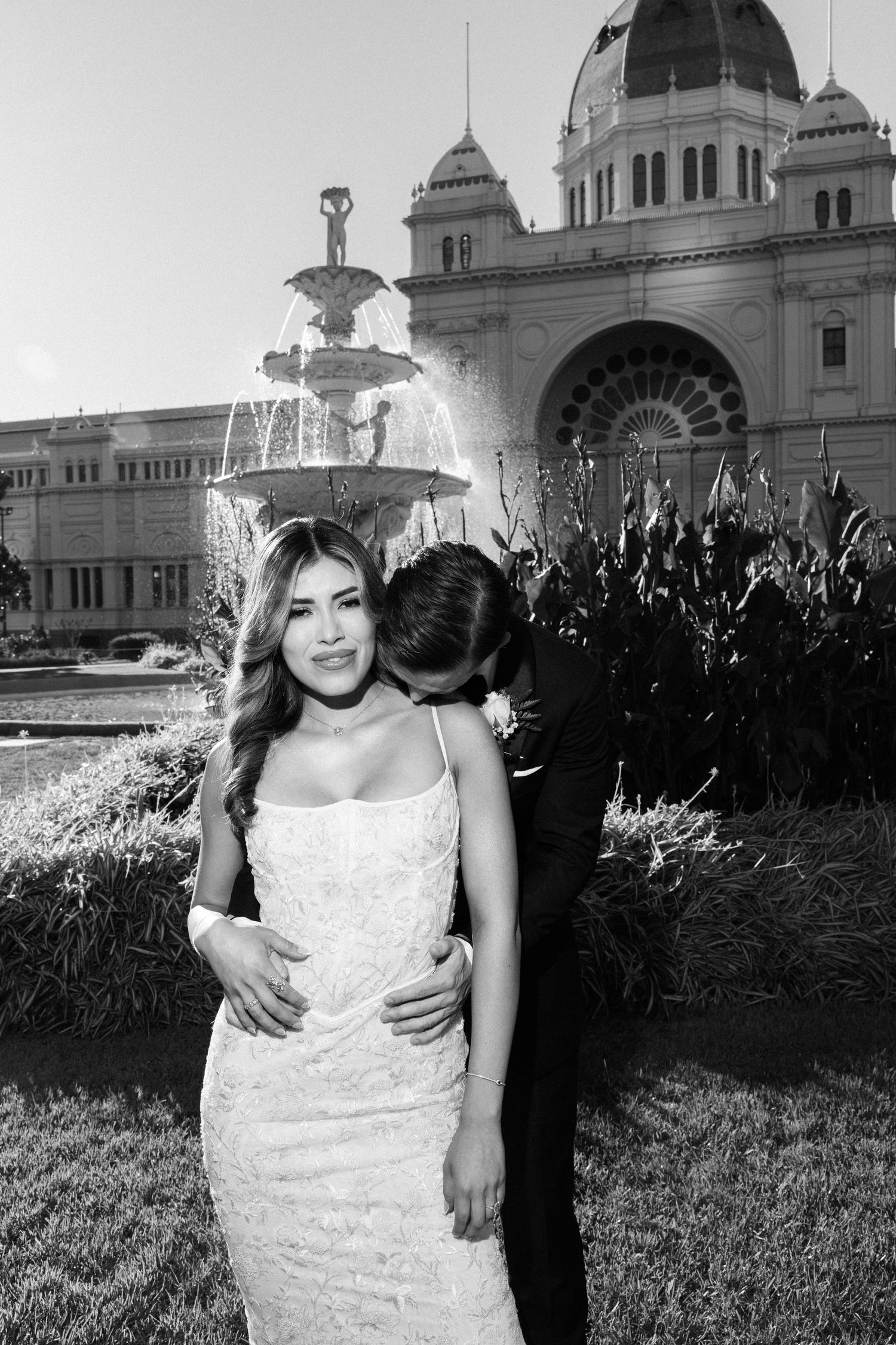 Black and white wedding photo of a couple embracing in front of a grand fountain and historic building.