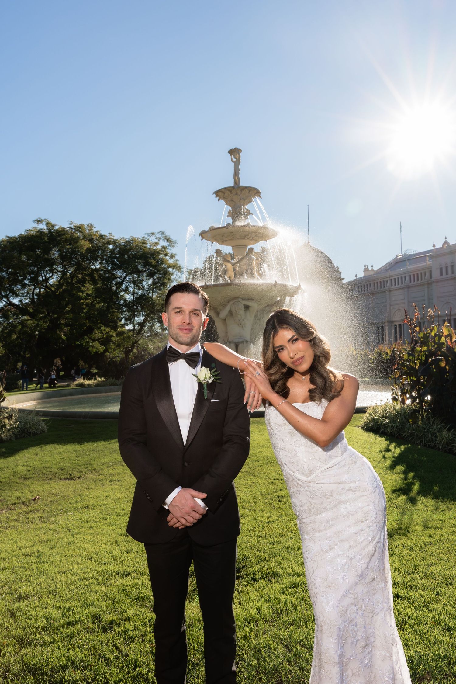 Wedding couple posing by an ornate fountain in sunlight, bride in lace gown, groom in black tuxedo.