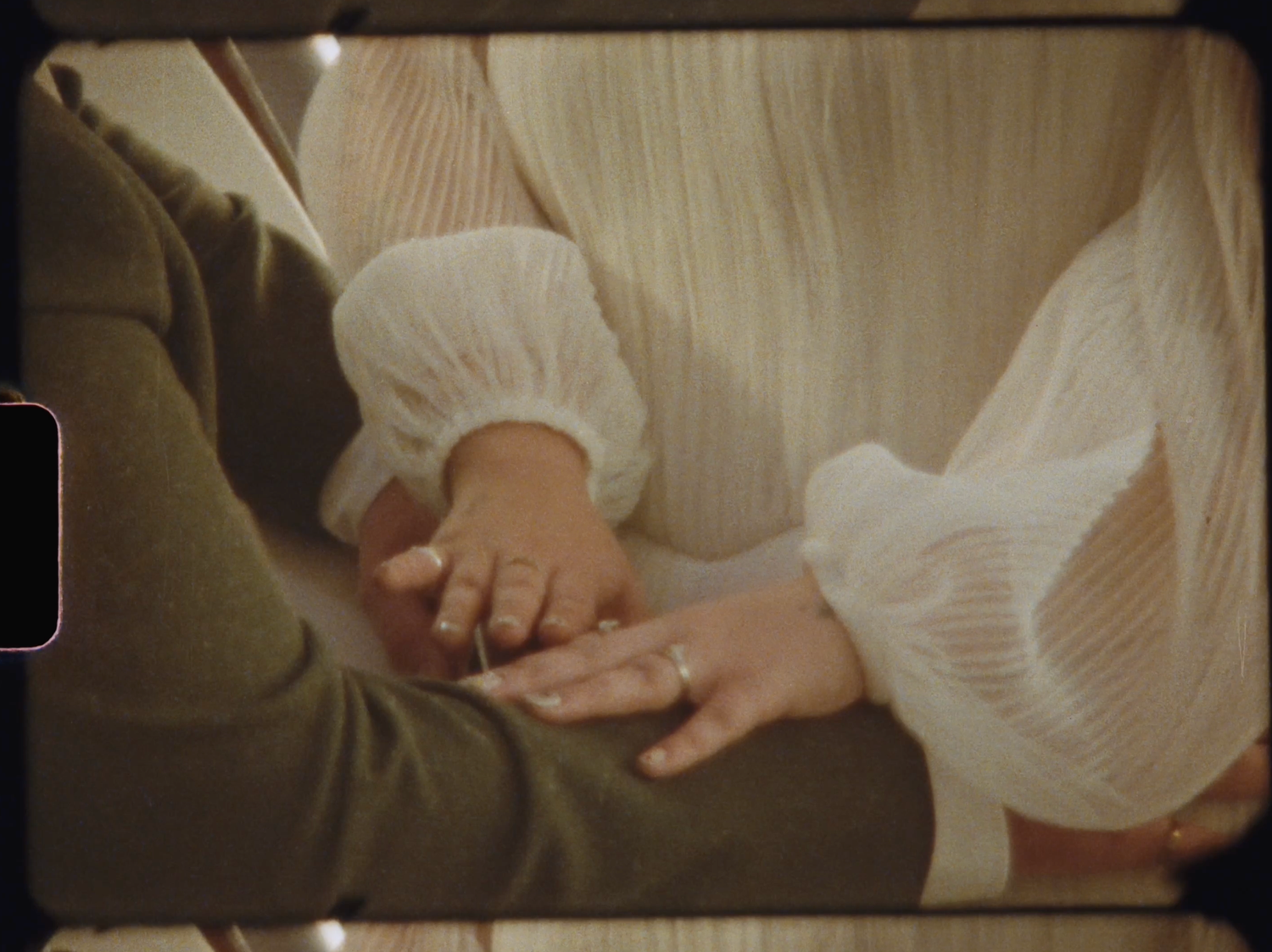 Close-up of two people holding hands, one wearing a diamond ring, dressed in olive and white pleated fabric.
