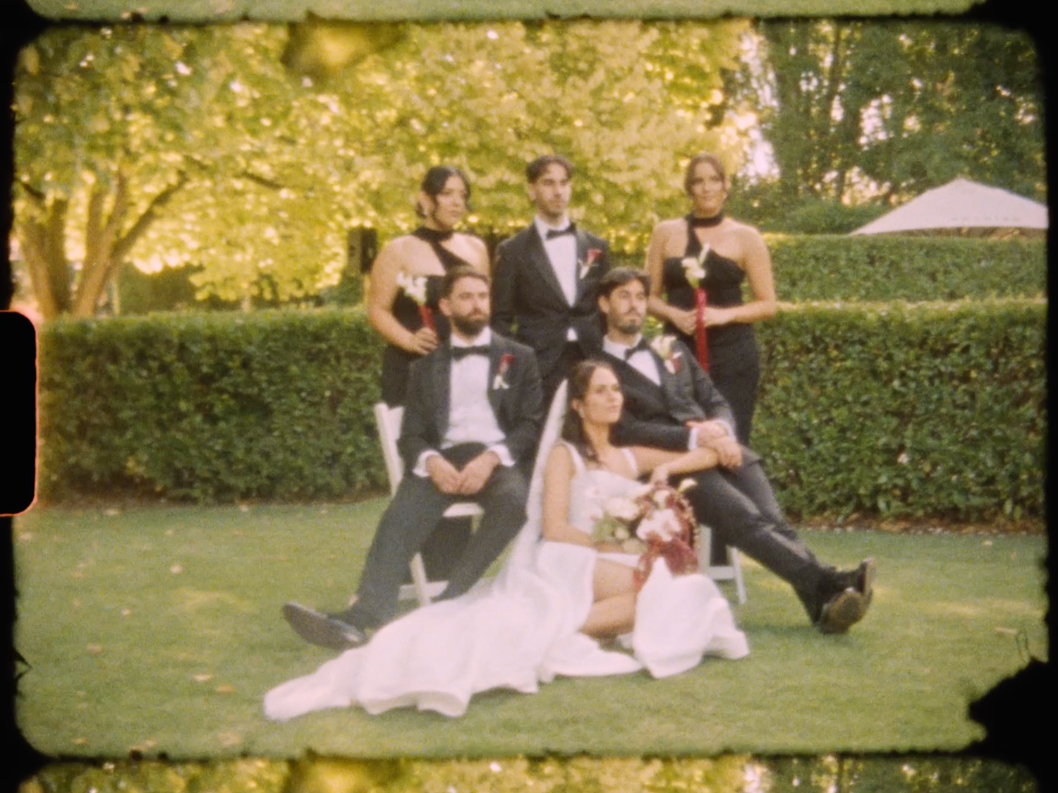 Wedding party posing in garden, bride in white gown with groom and bridesmaids in black attire on green lawn.