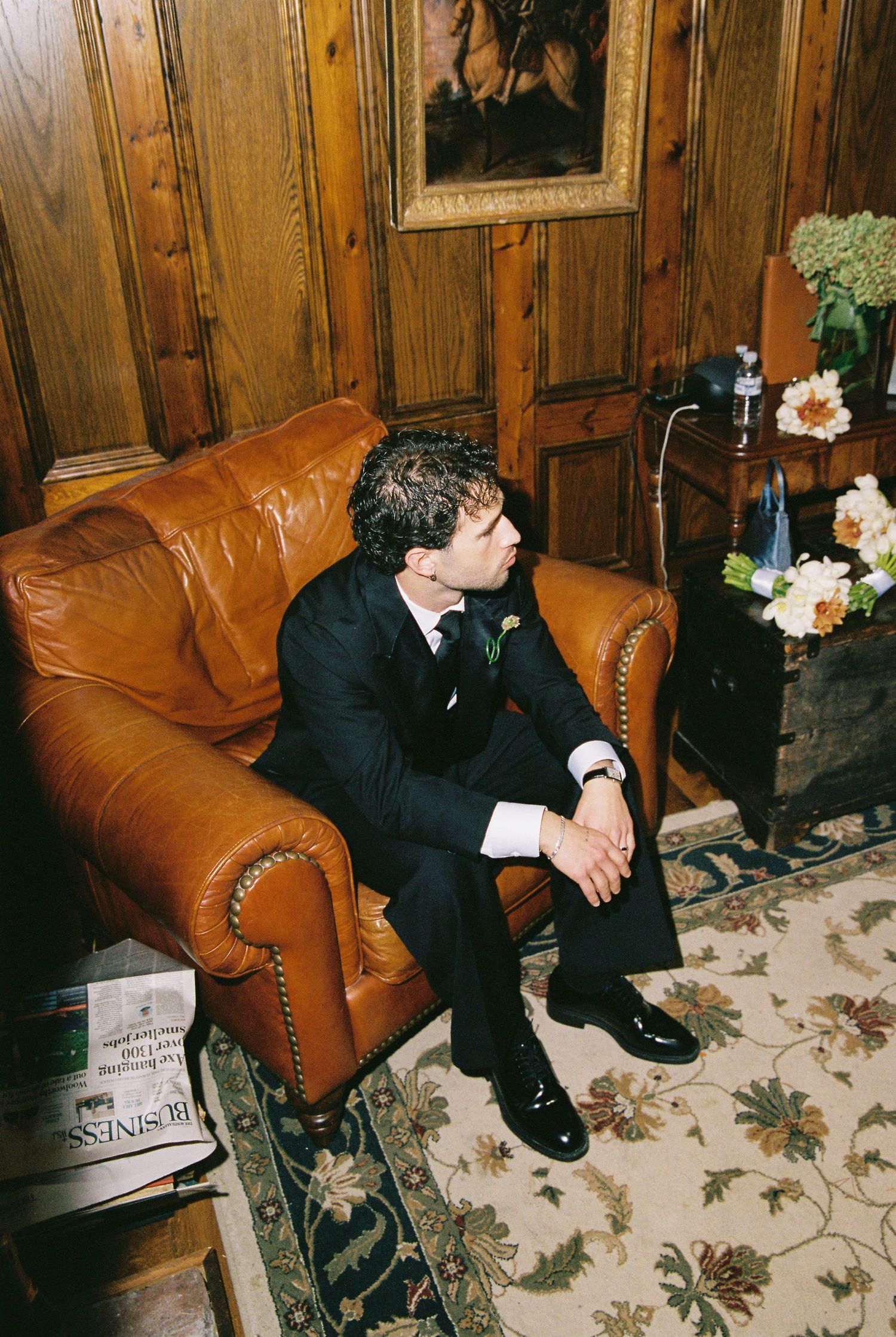 Groom in black suit sitting pensively in leather armchair in wood-paneled room before wedding ceremony.