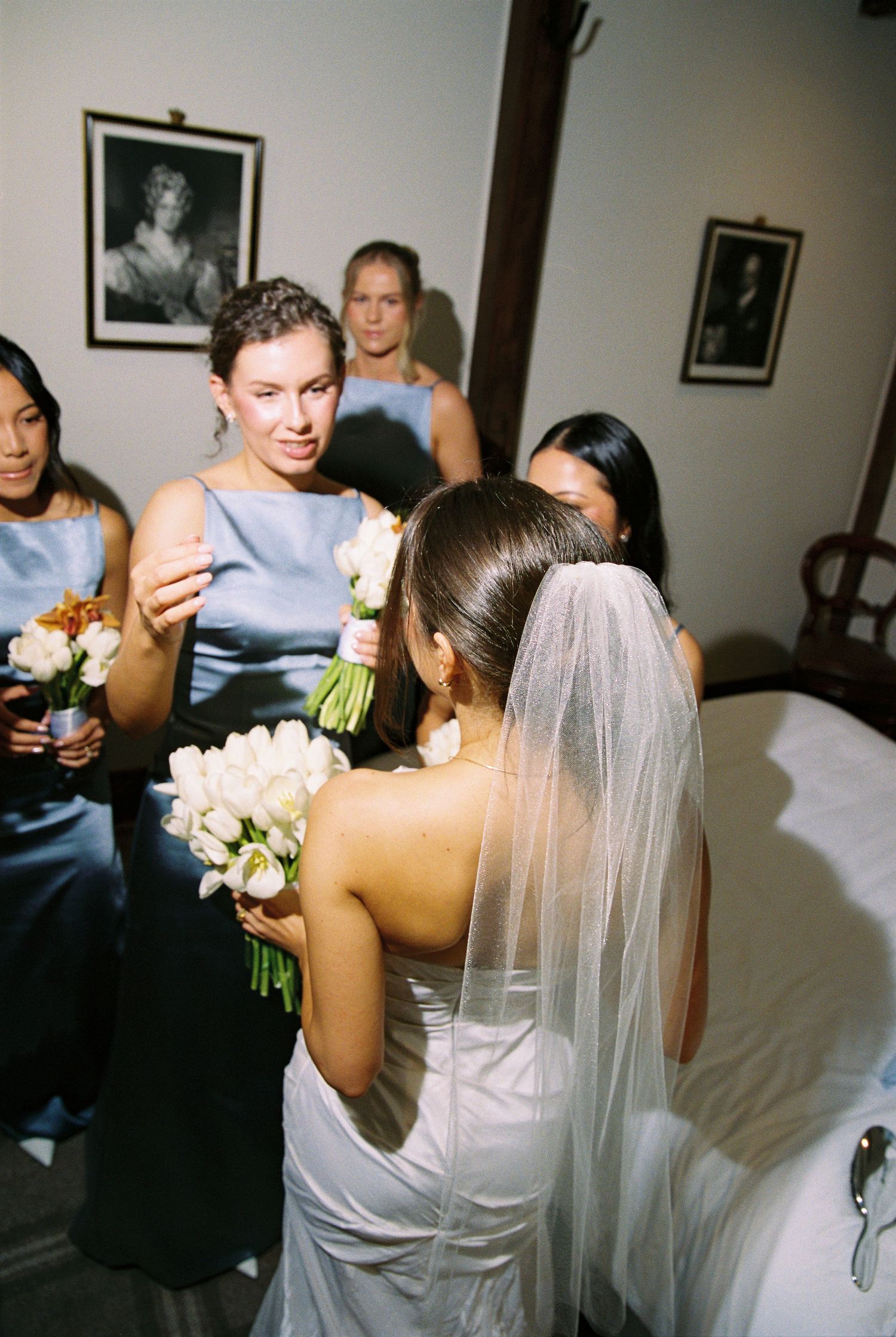 Bride in white gown and veil receives bouquet from bridesmaids in blue satin dresses during wedding preparations.