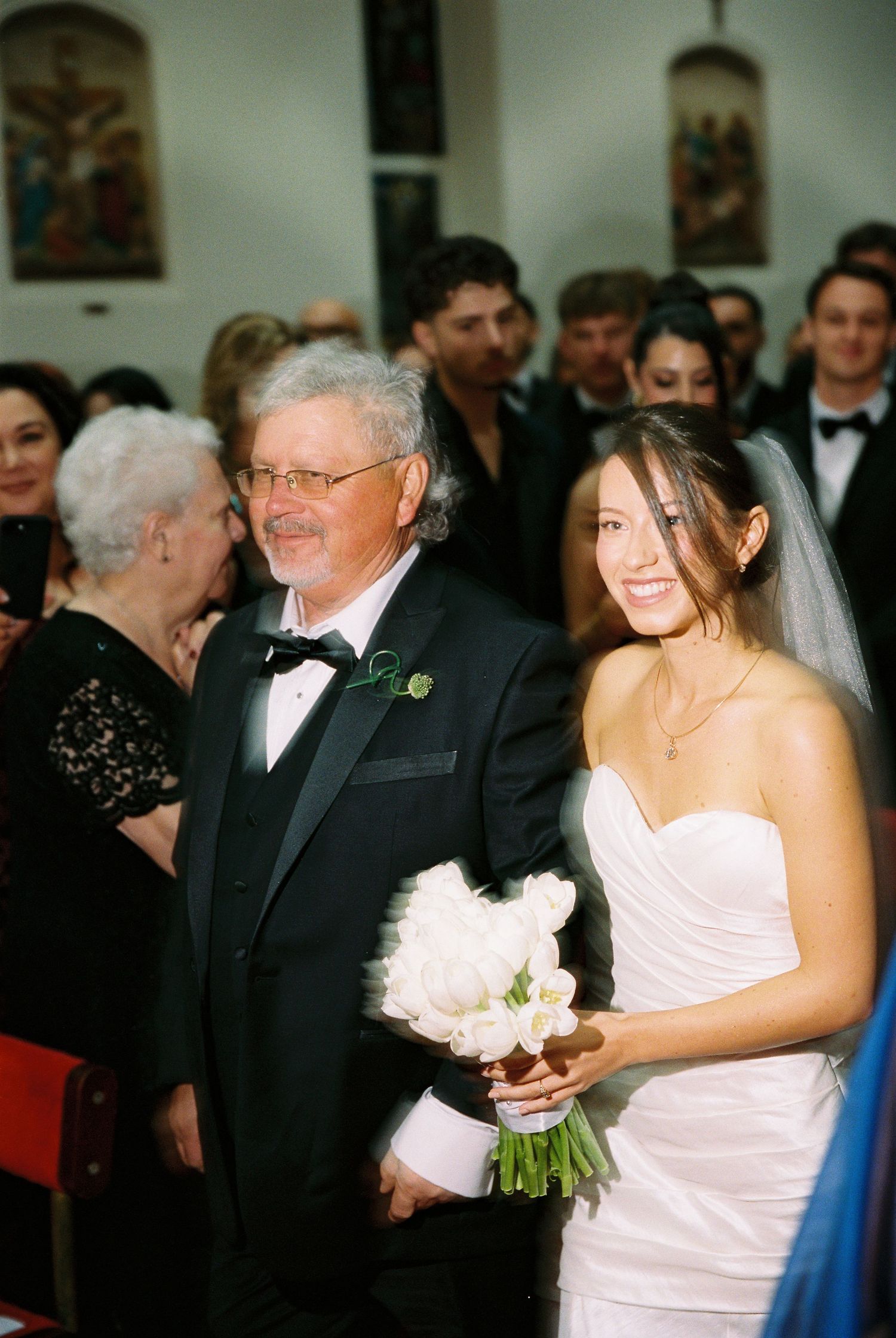 Father walks smiling bride down church aisle, bride holding white bouquet in strapless gown.
