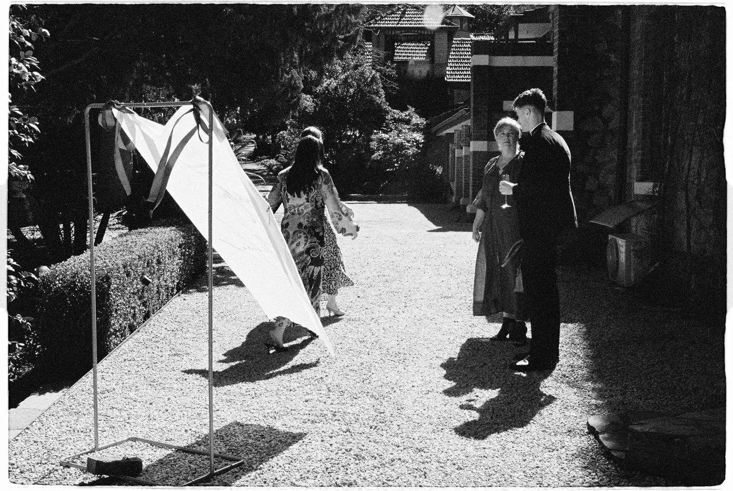 Black and white photo of people gathered in a sunlit garden path beside a white triangular sculpture.