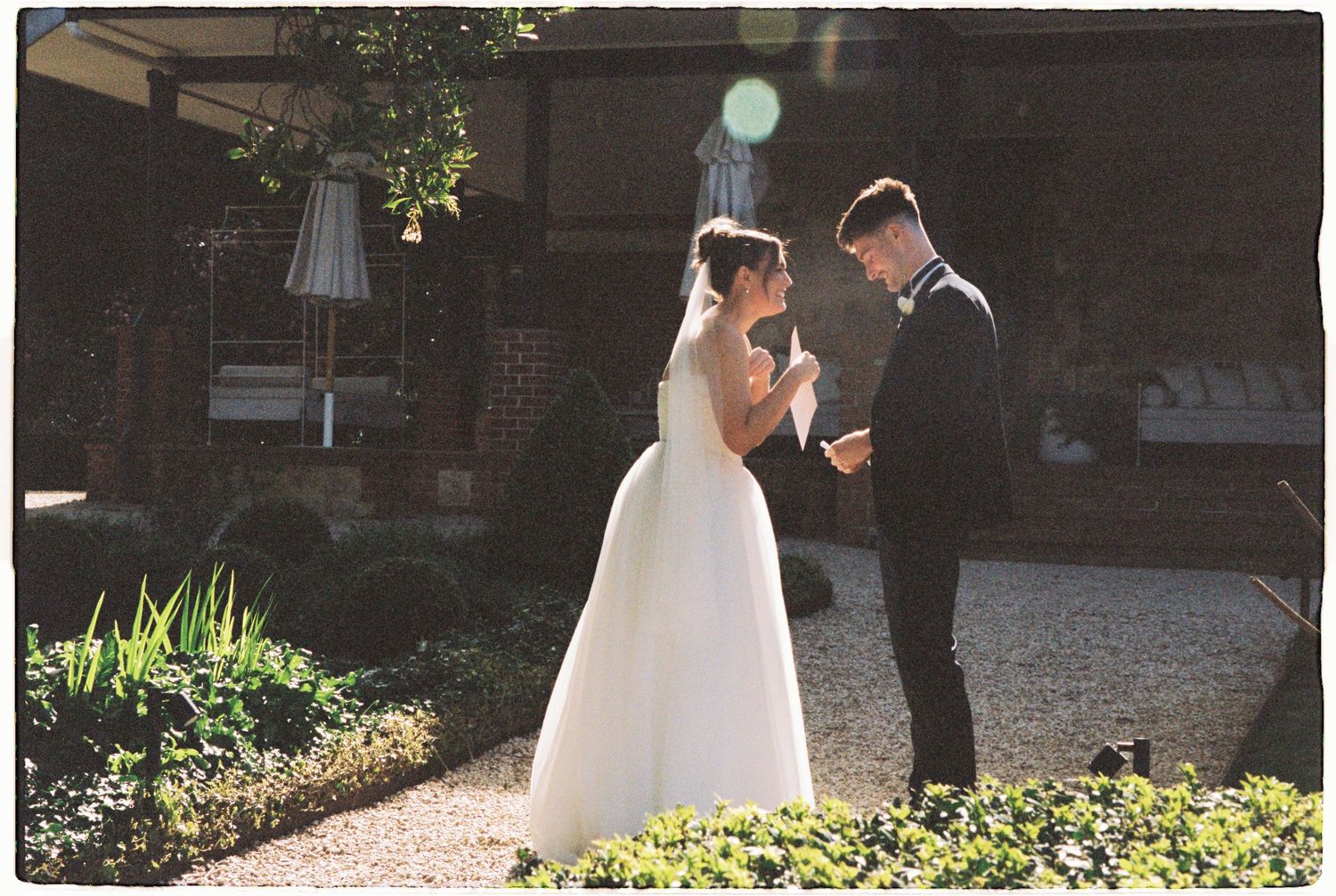 Bride and groom share a tender moment in a sunlit garden outside a brick venue.