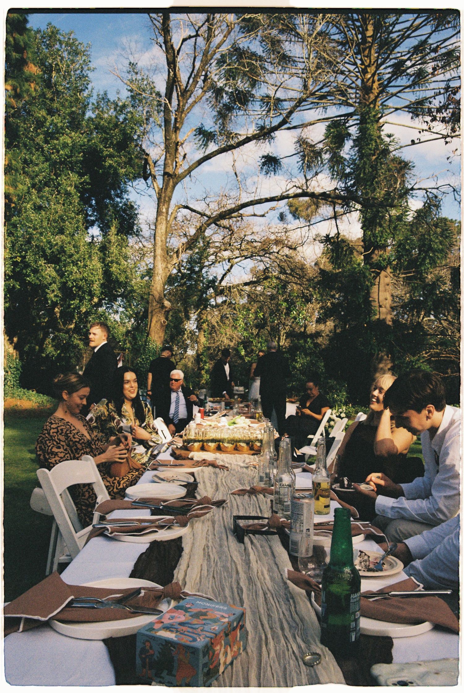 Guests seated at a long decorated outdoor wedding reception table surrounded by lush trees.