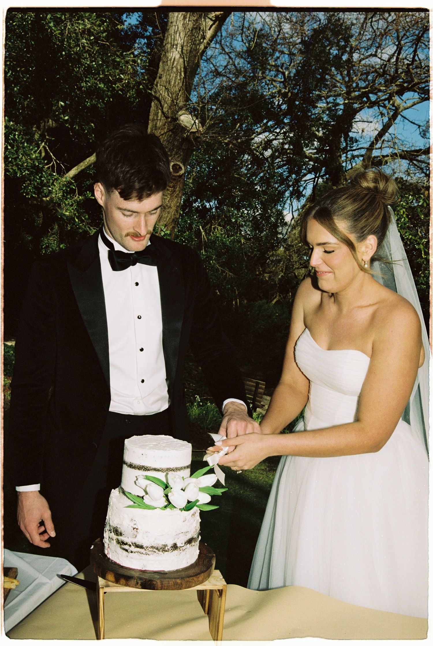Bride and groom cutting two-tier white wedding cake decorated with flowers outdoors.