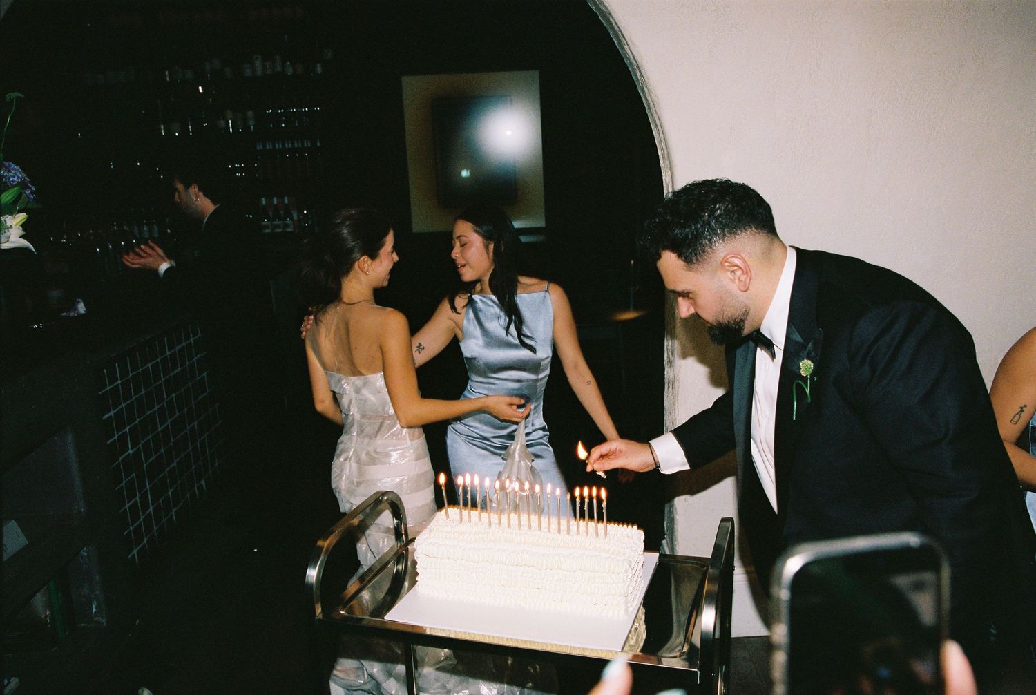 Guests gather around a white wedding cake with lit candles at an elegant evening reception.