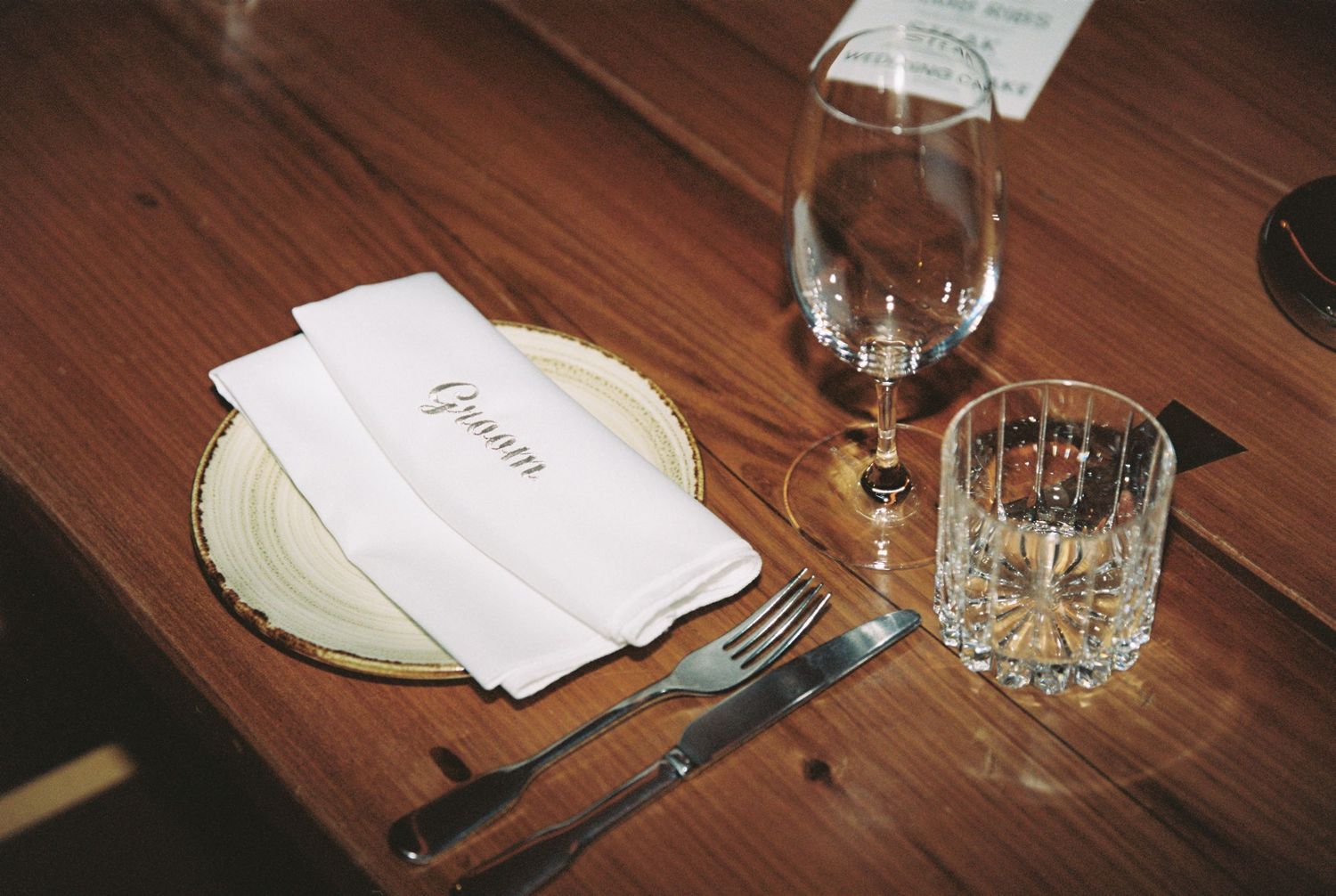 Elegant wedding place setting with Groom napkin, gold-rimmed plate, crystal glassware on wooden table.