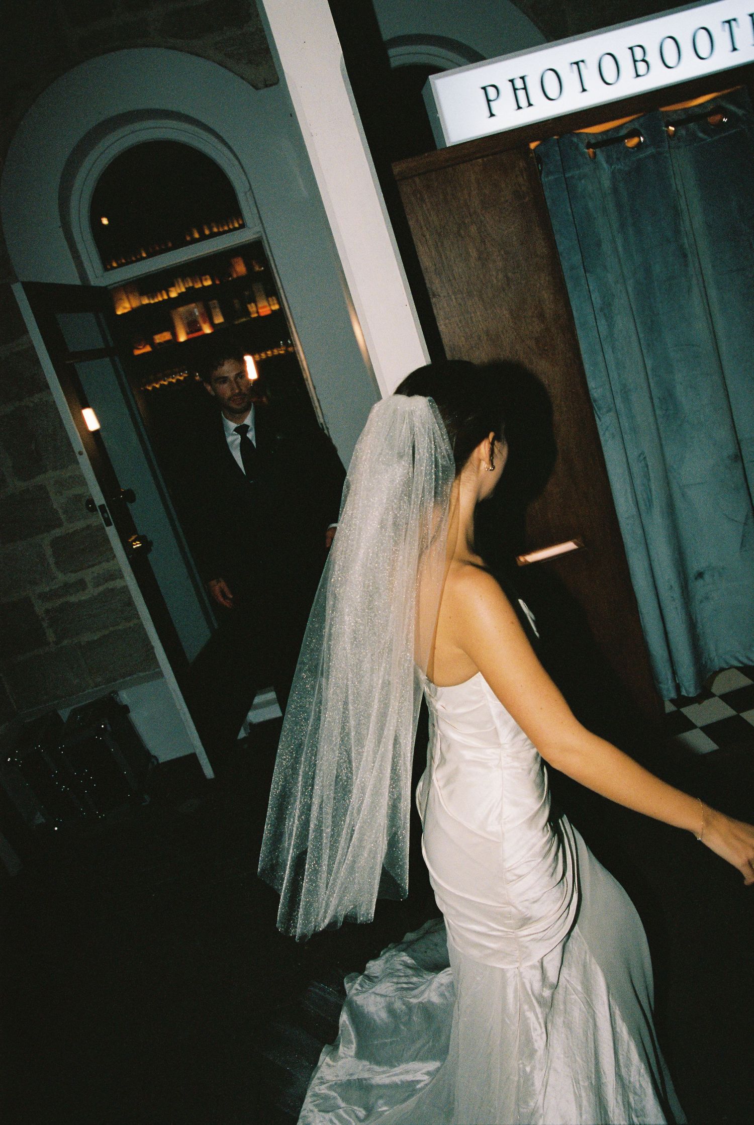 Bride in white gown and veil near a photobooth at a wedding reception with groom in background.