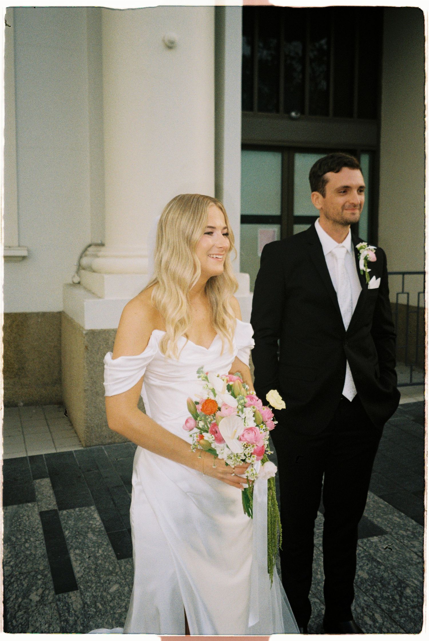 Smiling blonde bride in elegant white gown holding colorful bouquet with groom in black suit behind.