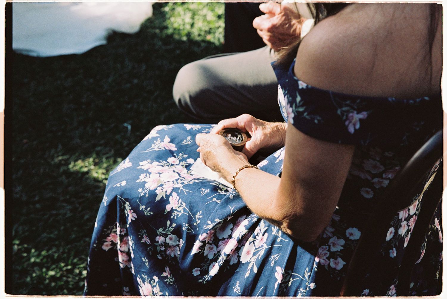 Guest in blue floral dress holds small object during outdoor wedding ceremony.