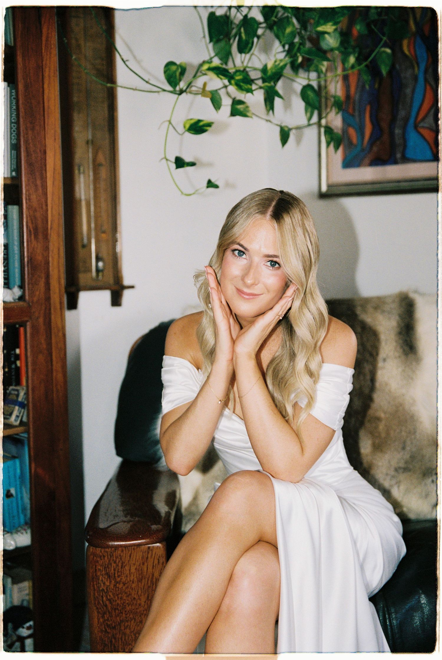 Beautiful blonde bride in white off-shoulder dress posing with hands on cheeks indoors.