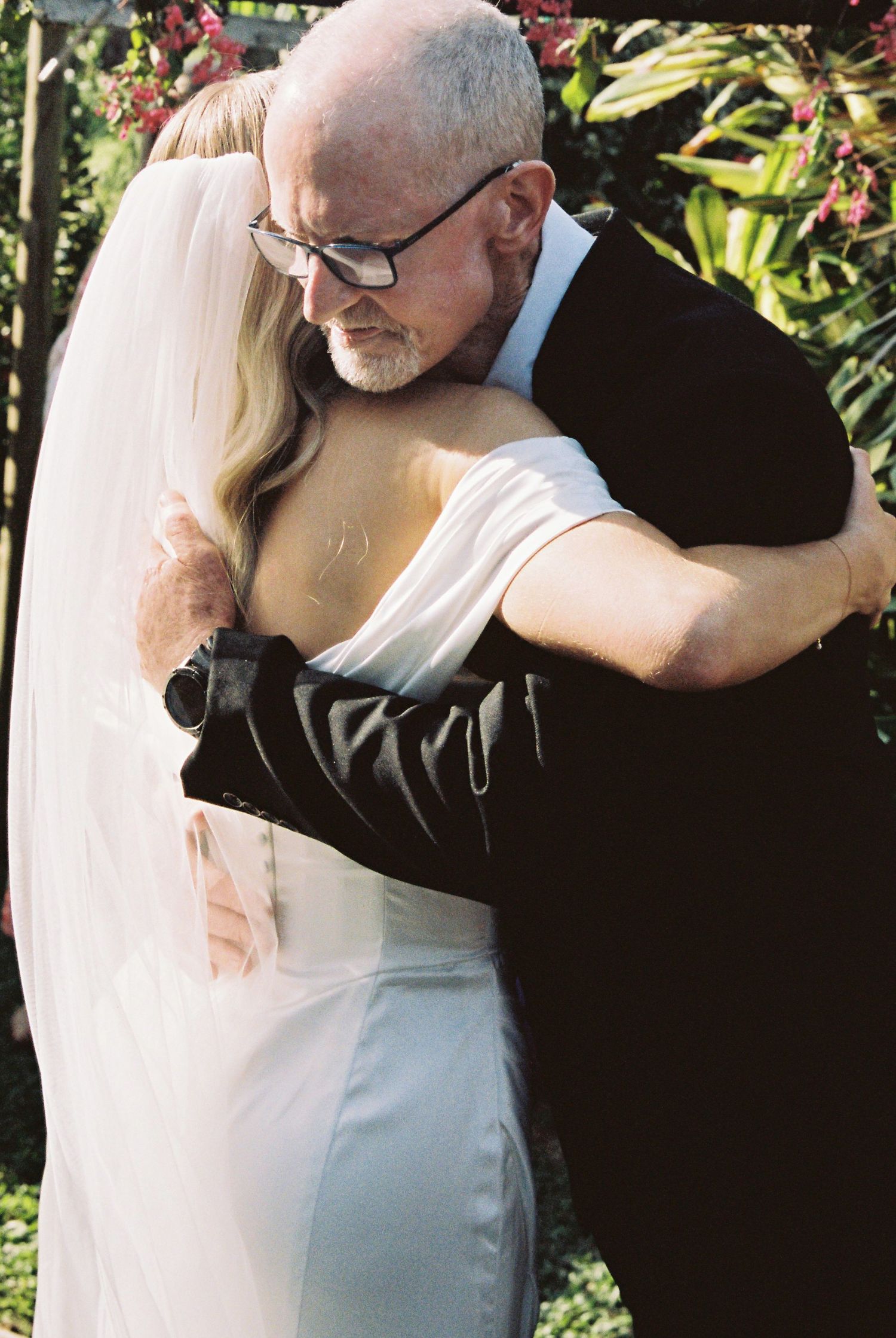 Emotional father-daughter embrace at outdoor wedding ceremony, bride in white off-shoulder gown and veil.