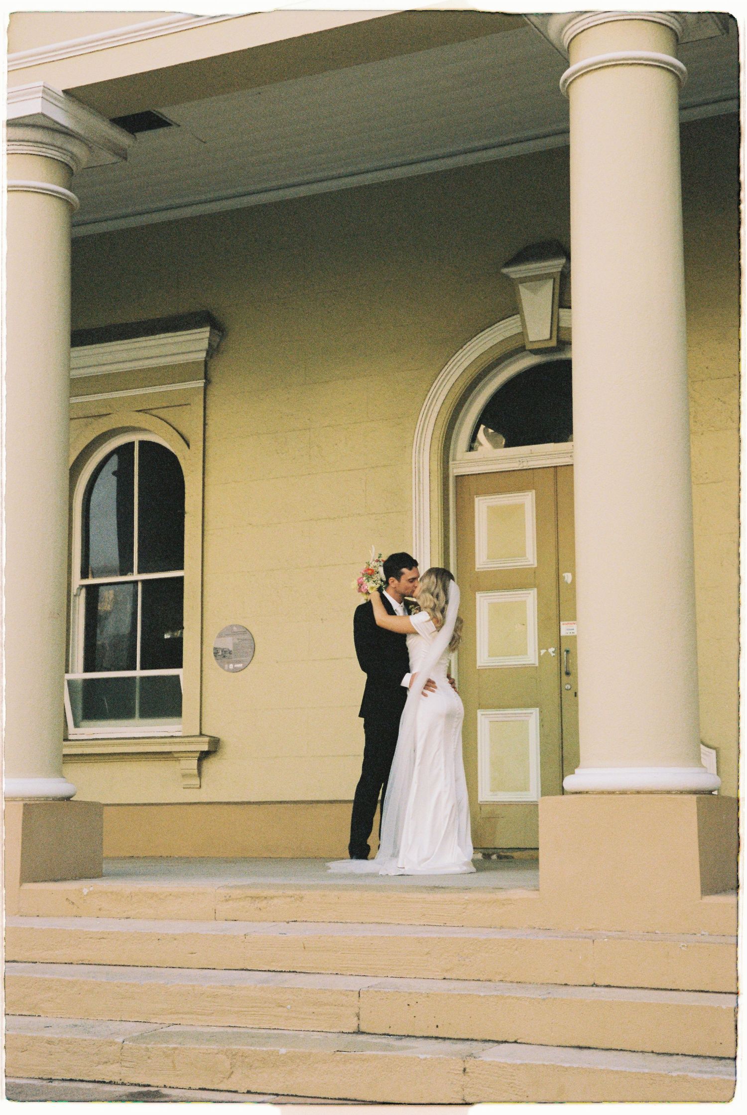 Bride and groom share a kiss on steps of yellow heritage building with white columns.