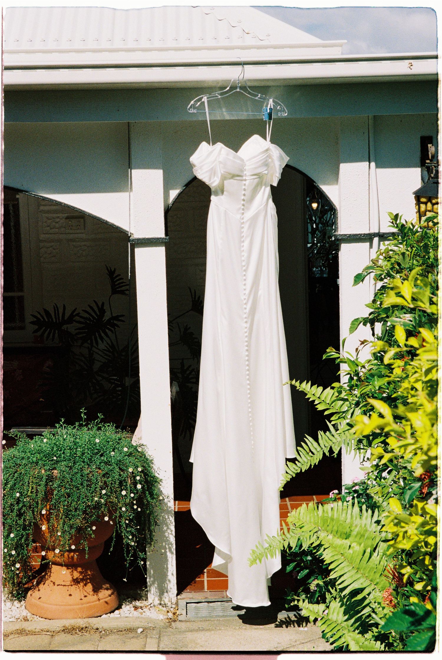 White wedding dress hanging on hanger outside house surrounded by lush green garden plants.