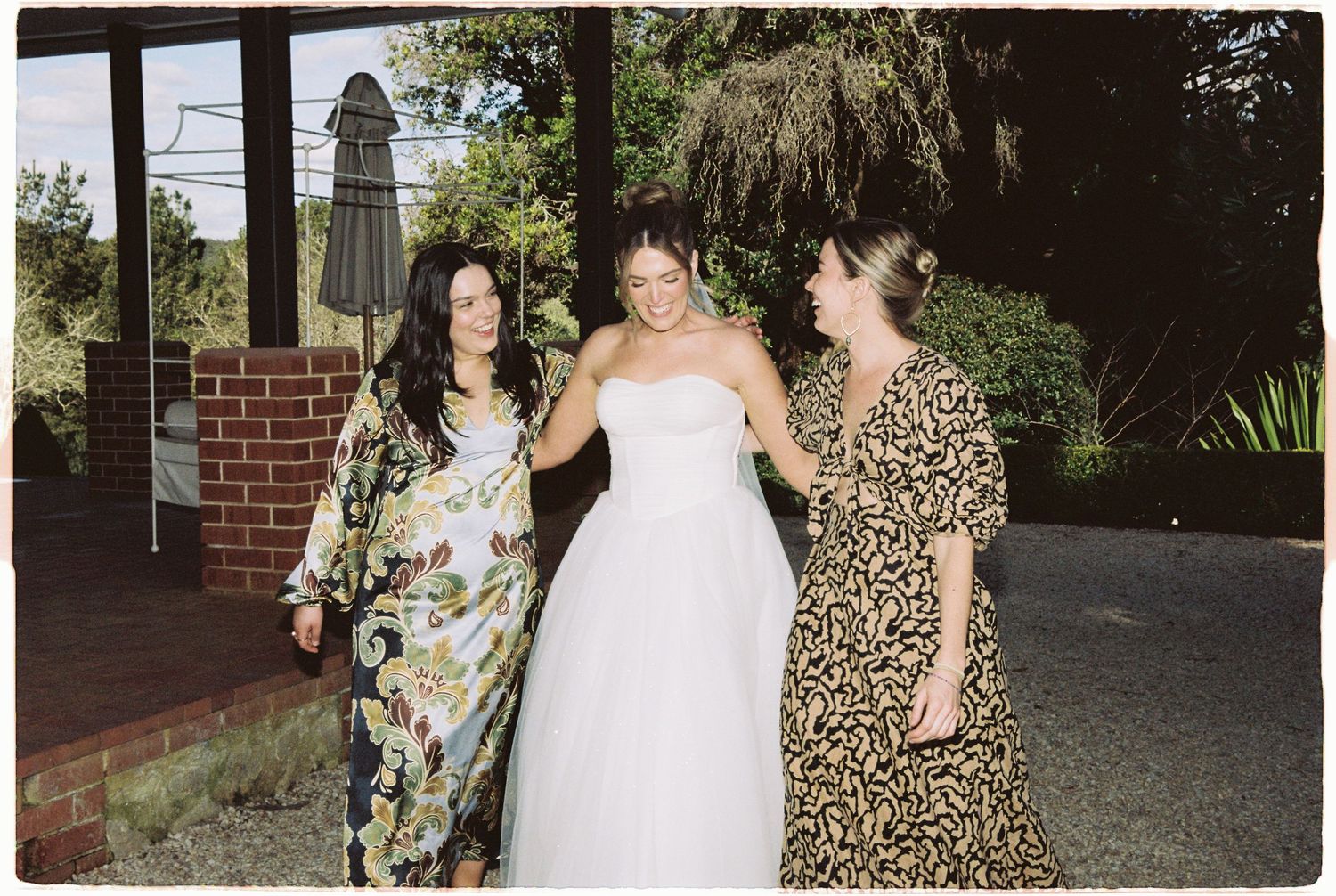Bride in white strapless gown posing with two guests in floral dresses at outdoor wedding.
