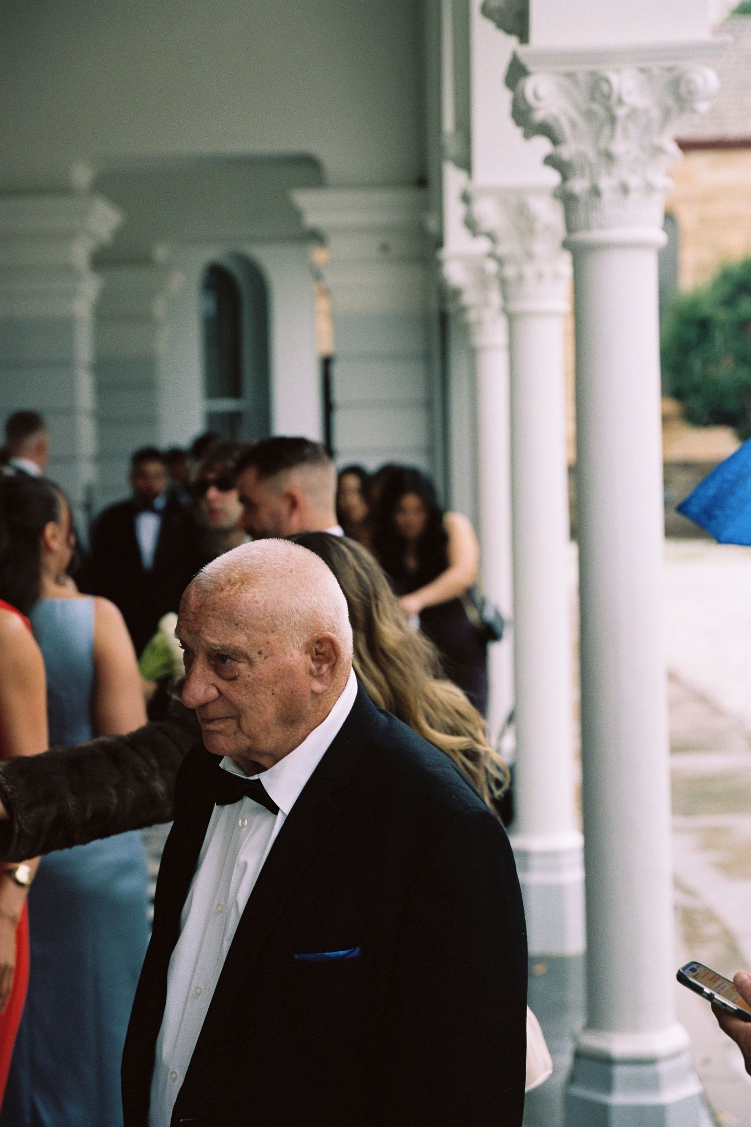 Elderly man in black tuxedo with bow tie stands among wedding guests at a columned venue.