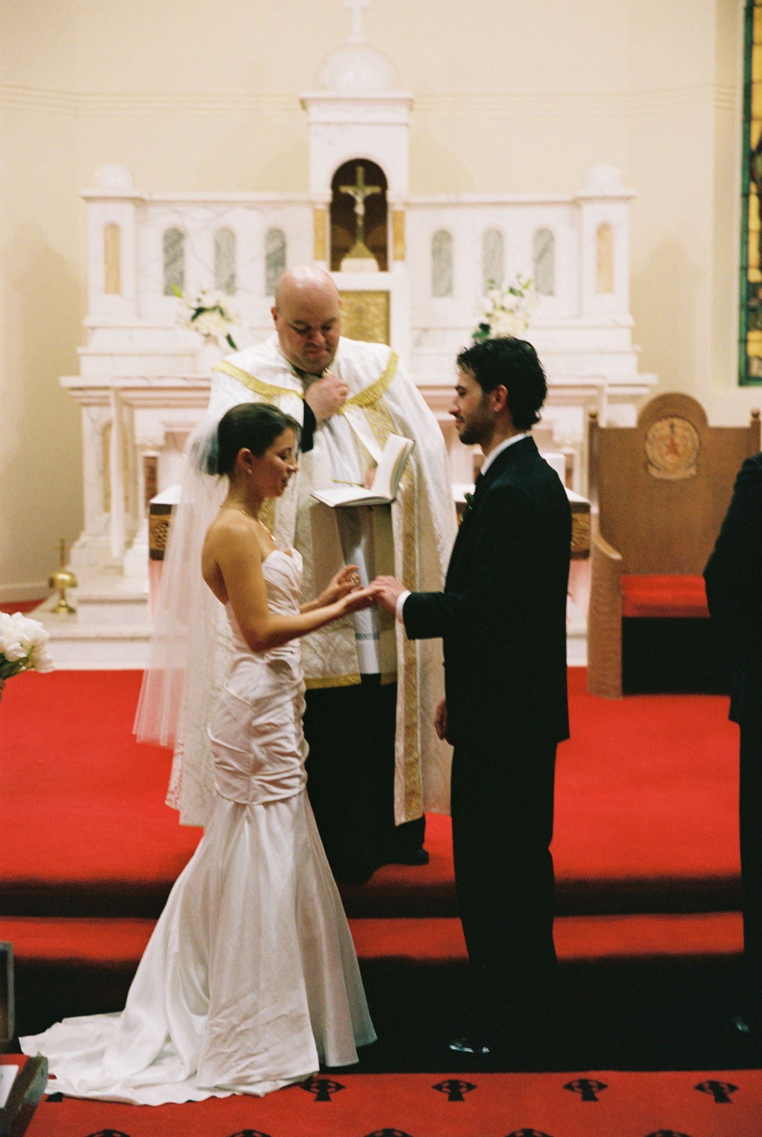 Bride and groom exchange vows before a priest at a traditional church wedding ceremony with red carpet and white altar.