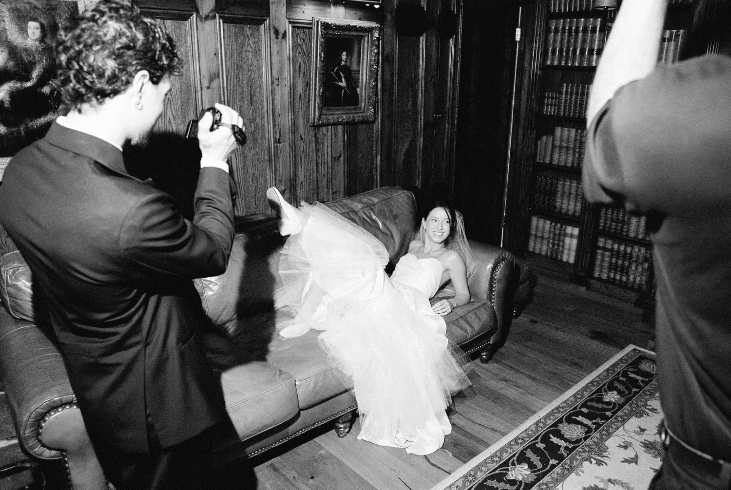 Groom photographing bride reclining on leather sofa in elegant wood-paneled library.