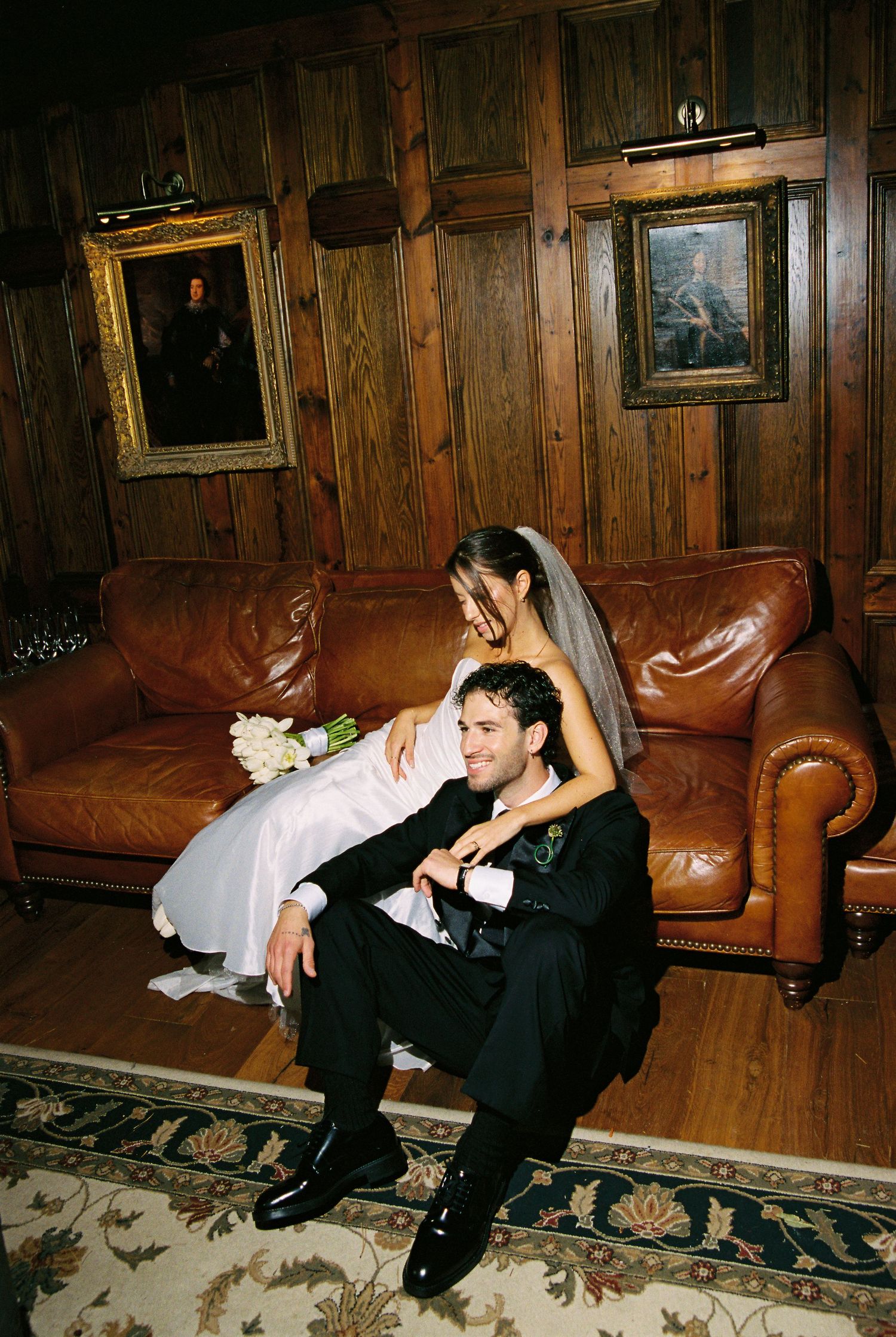 Bride and groom sitting together on a brown leather couch in a wood-paneled room with framed portraits.