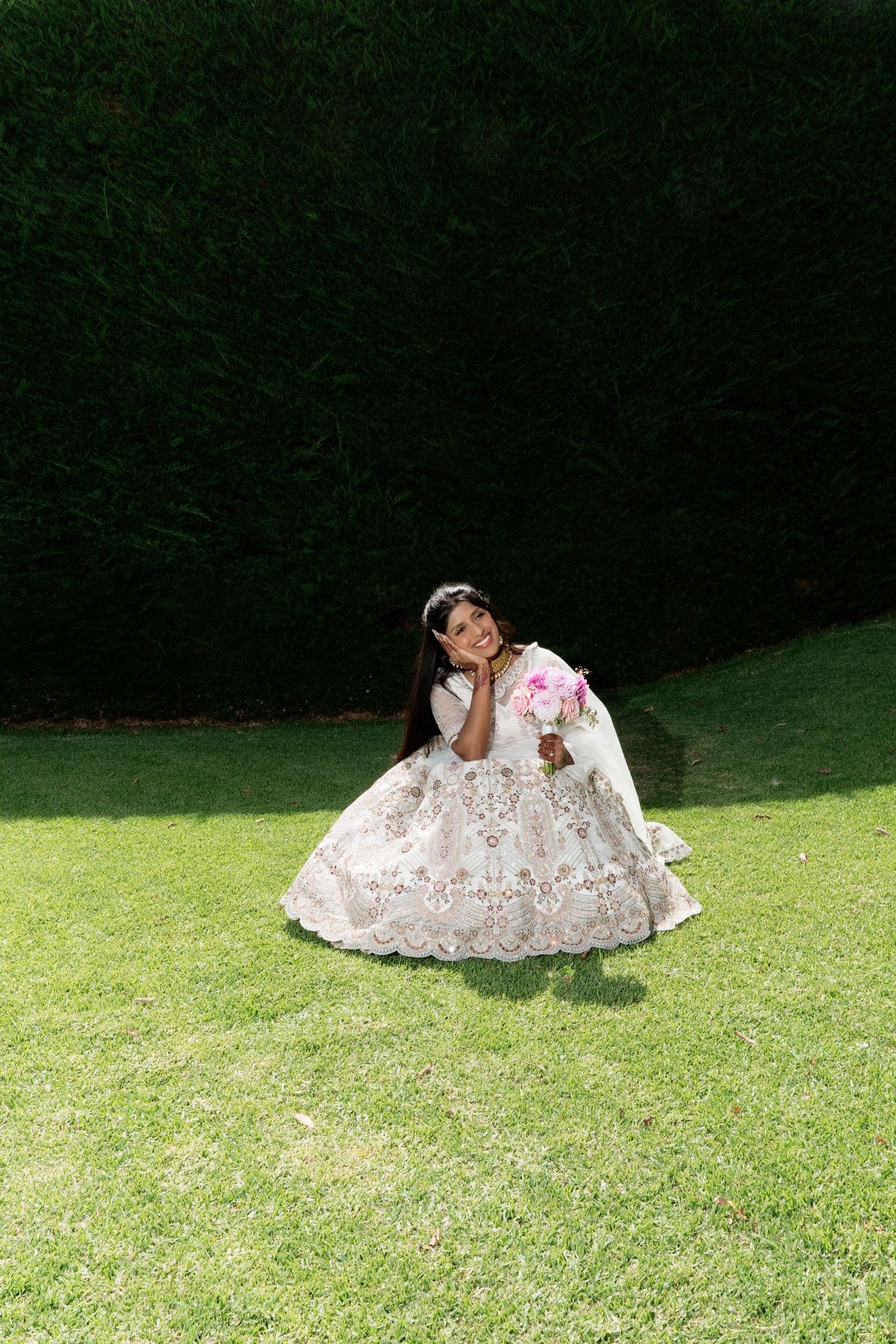Bride in ornate white embroidered lehenga sitting gracefully on green lawn holding pink bouquet on wedding day.