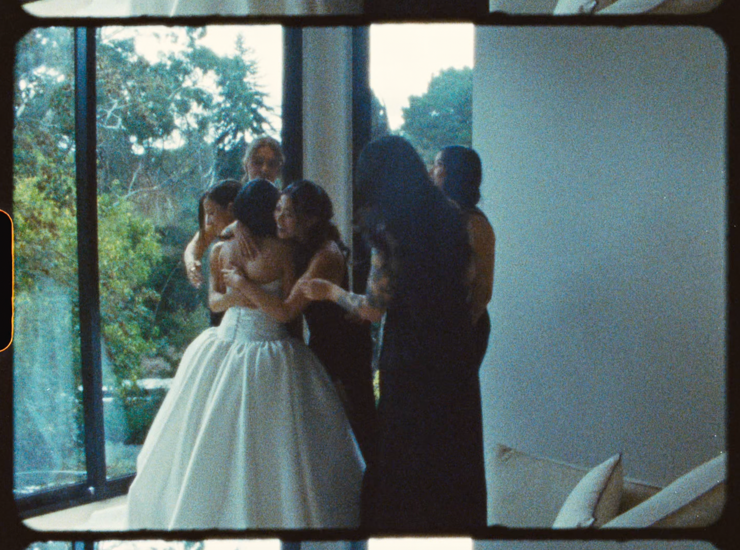 Bride in white ball gown embraces bridesmaids in black dresses in a modern glass-walled room.