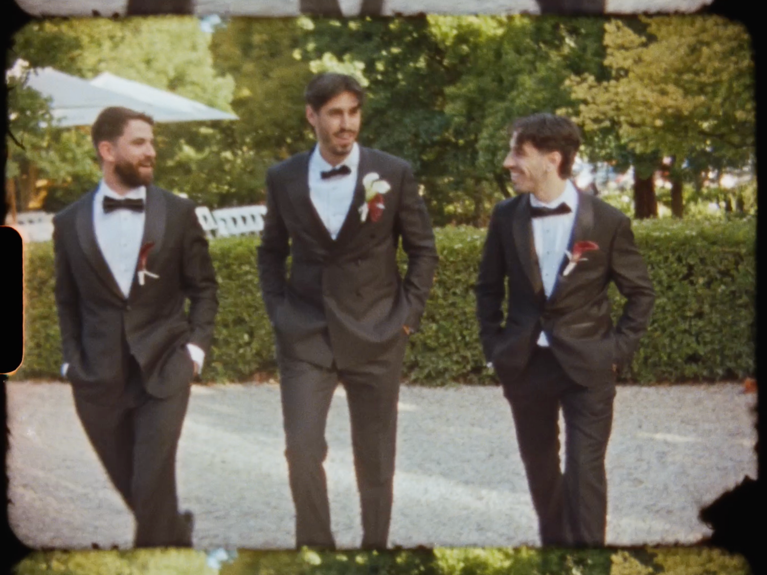 Three groomsmen in dark suits with bow ties and boutonnieres walking outdoors at a wedding.