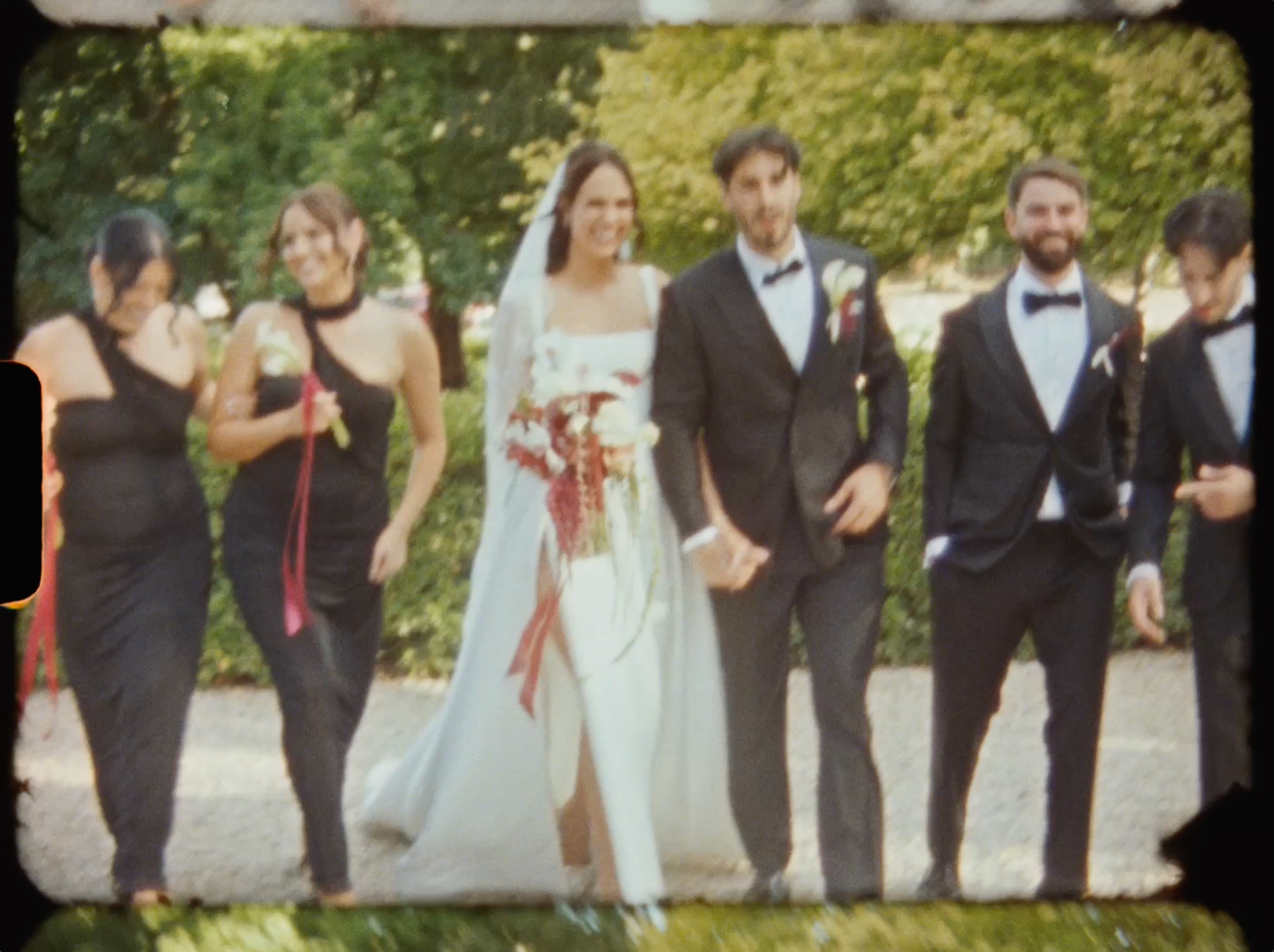 Wedding party walks together outdoors, bride in white gown flanked by bridesmaids in black and groomsmen in tuxedos.
